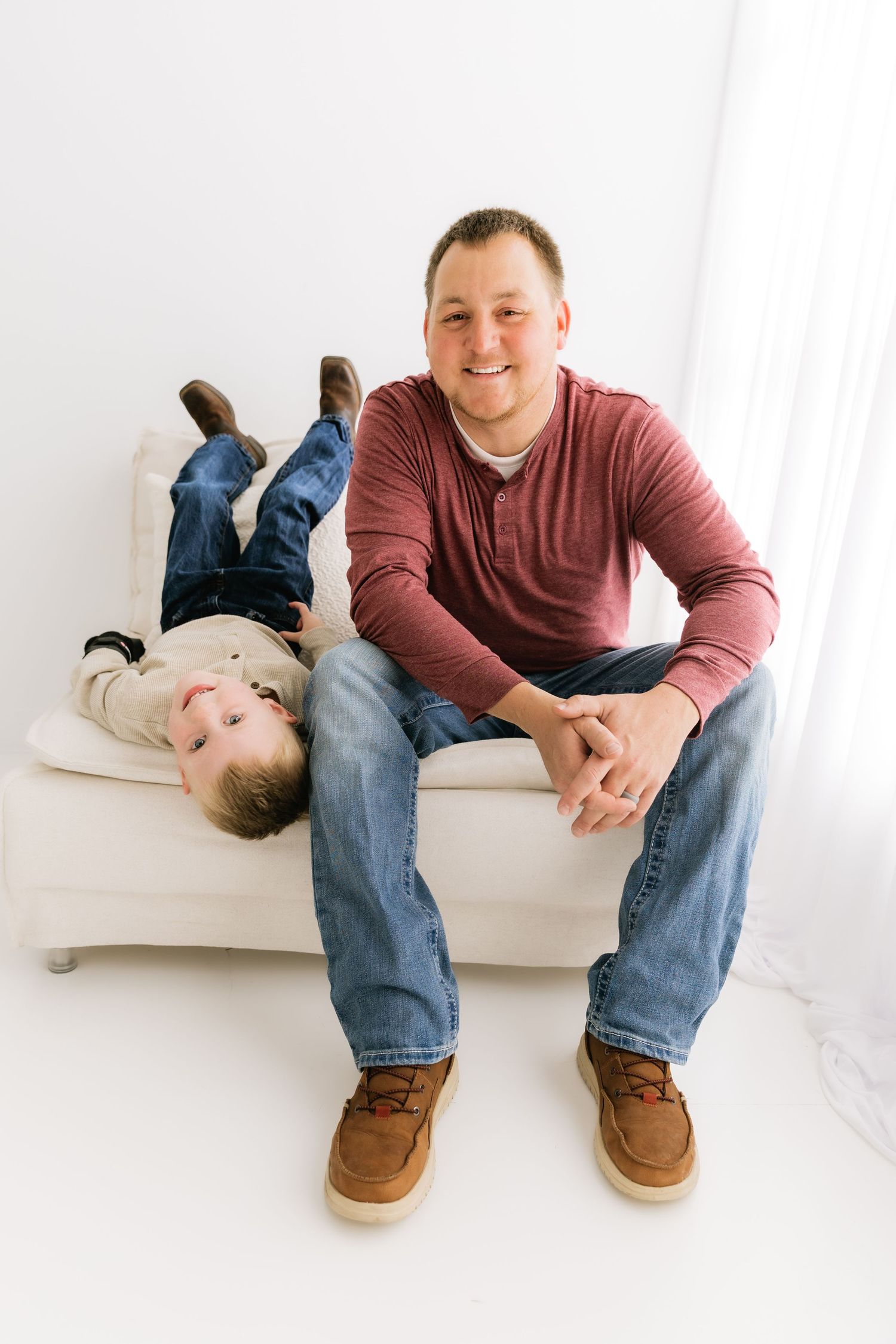 Smiling father sits on white bench with young son laying upside down beside him indoors.