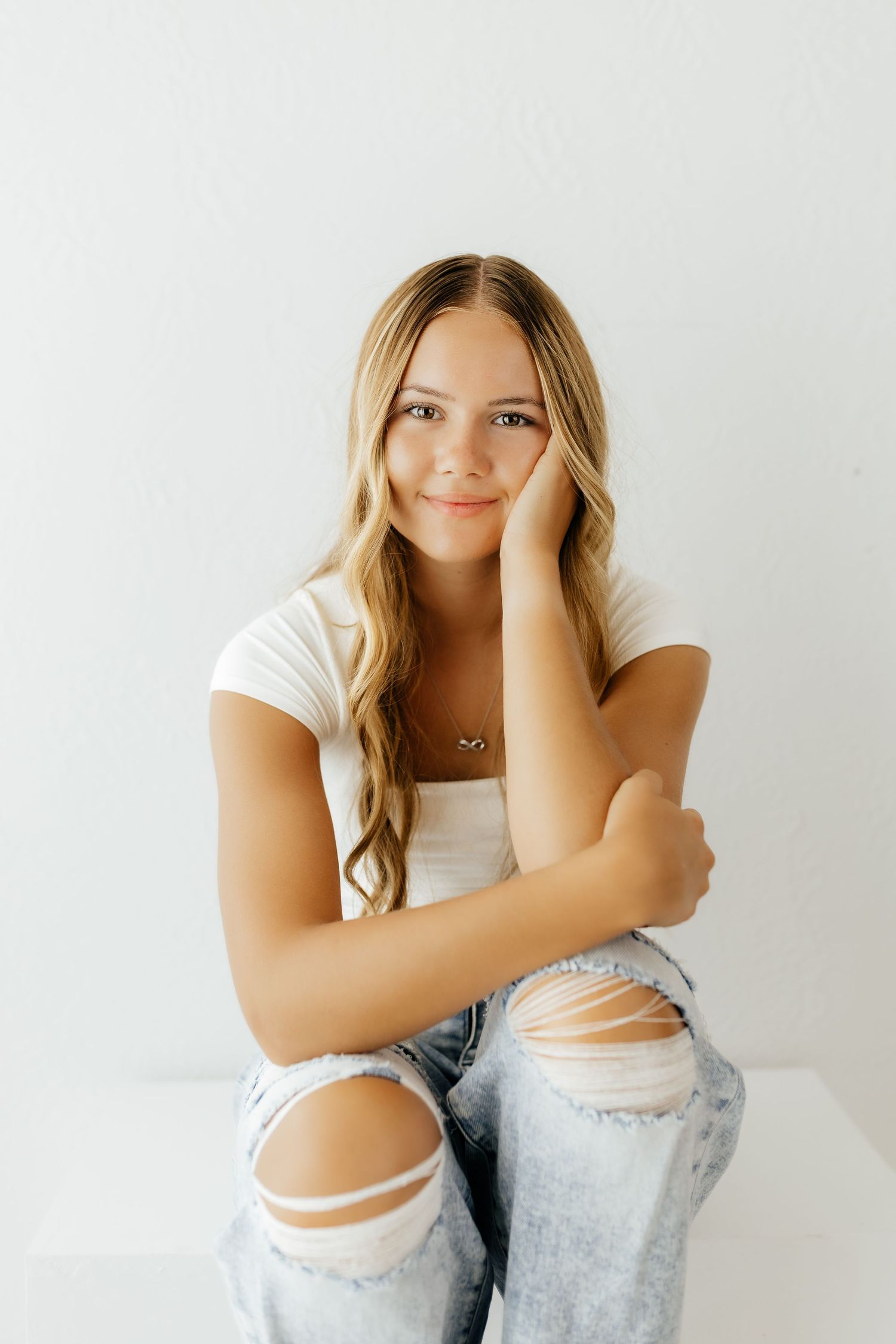 A smiling teenage girl with long blonde hair poses in ripped jeans and white top against a white wall.