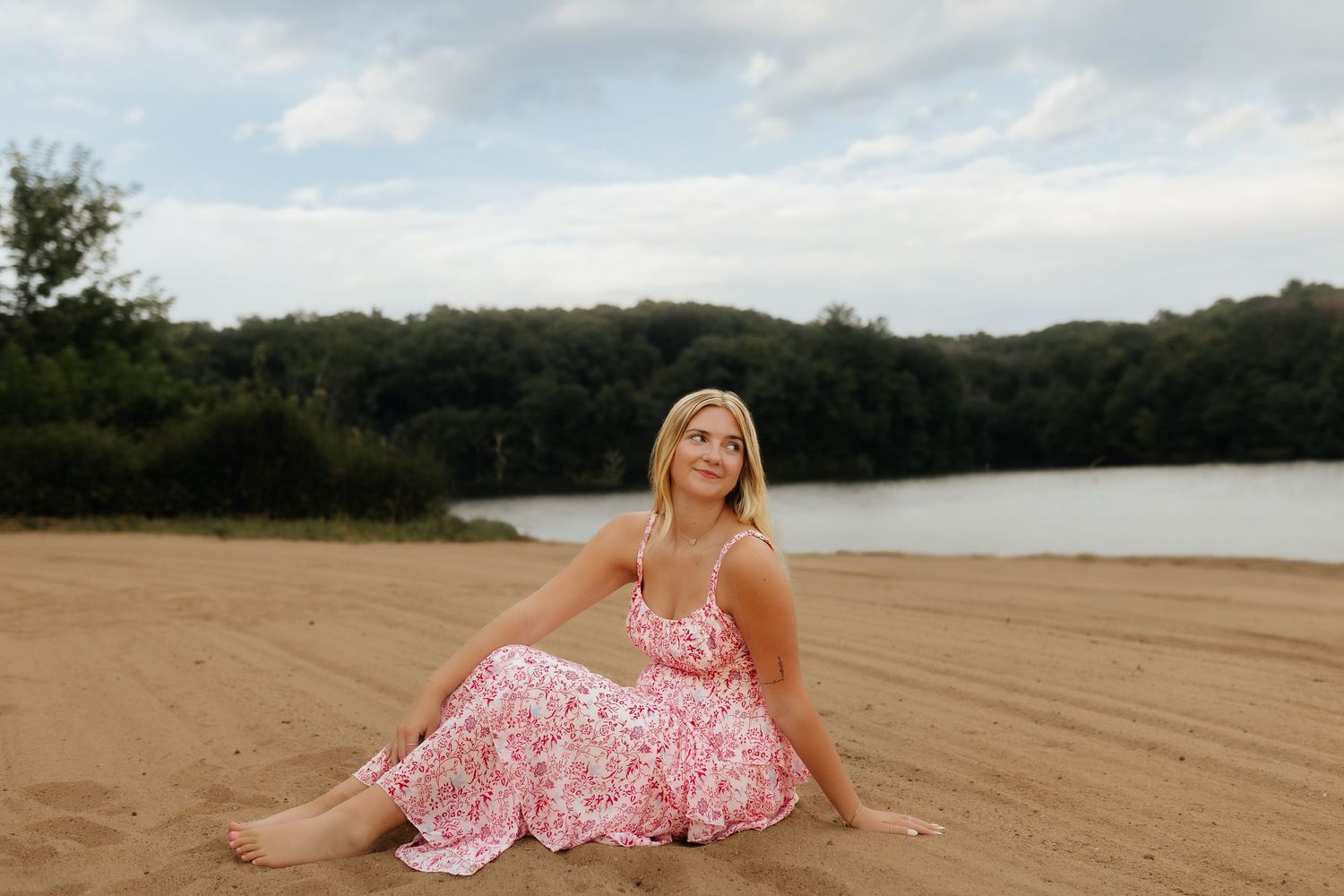 Blonde woman in pink floral dress sitting barefoot on sandy beach with lake and trees behind.