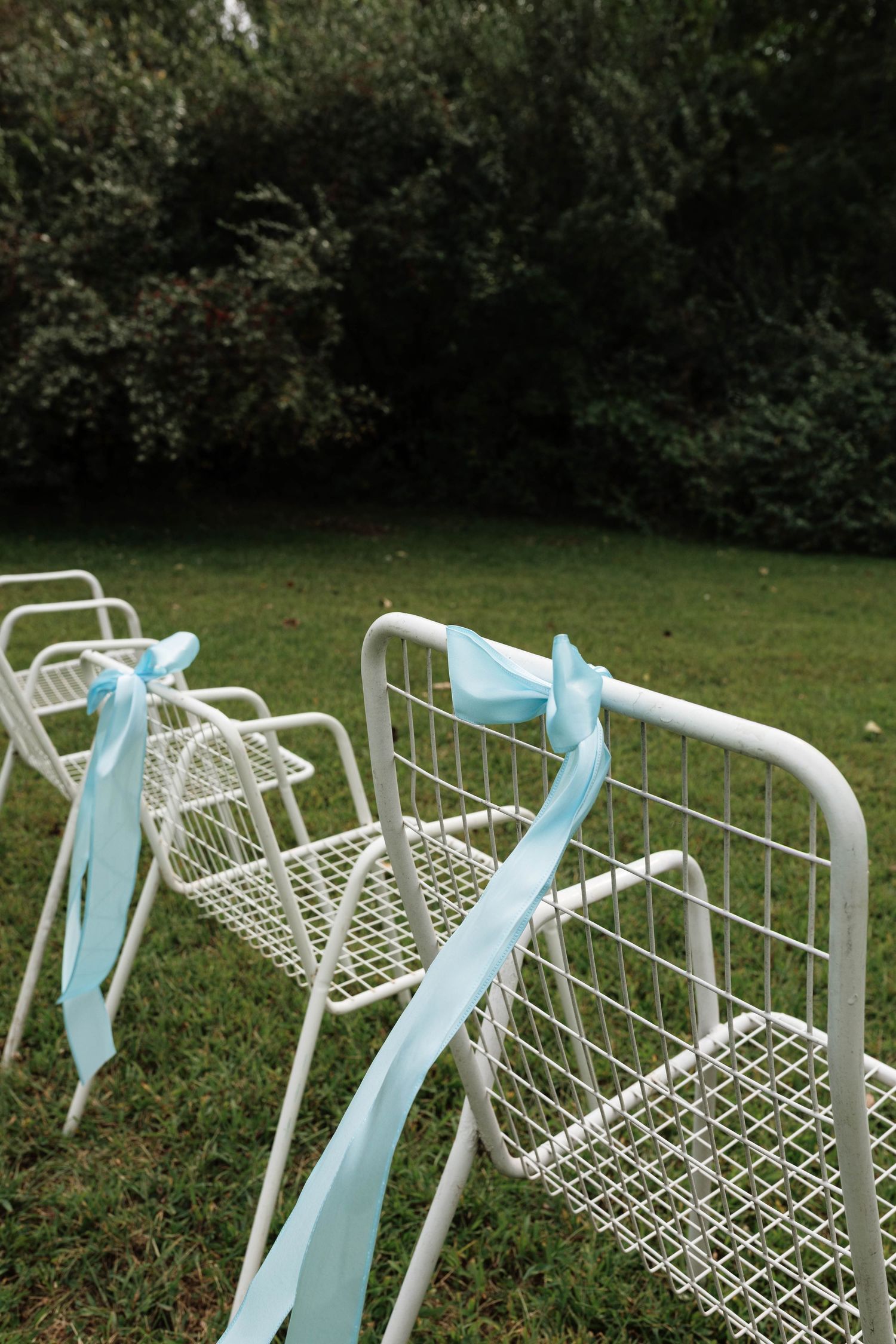 White metal chairs decorated with light blue ribbon bows arranged on green grass for outdoor wedding.