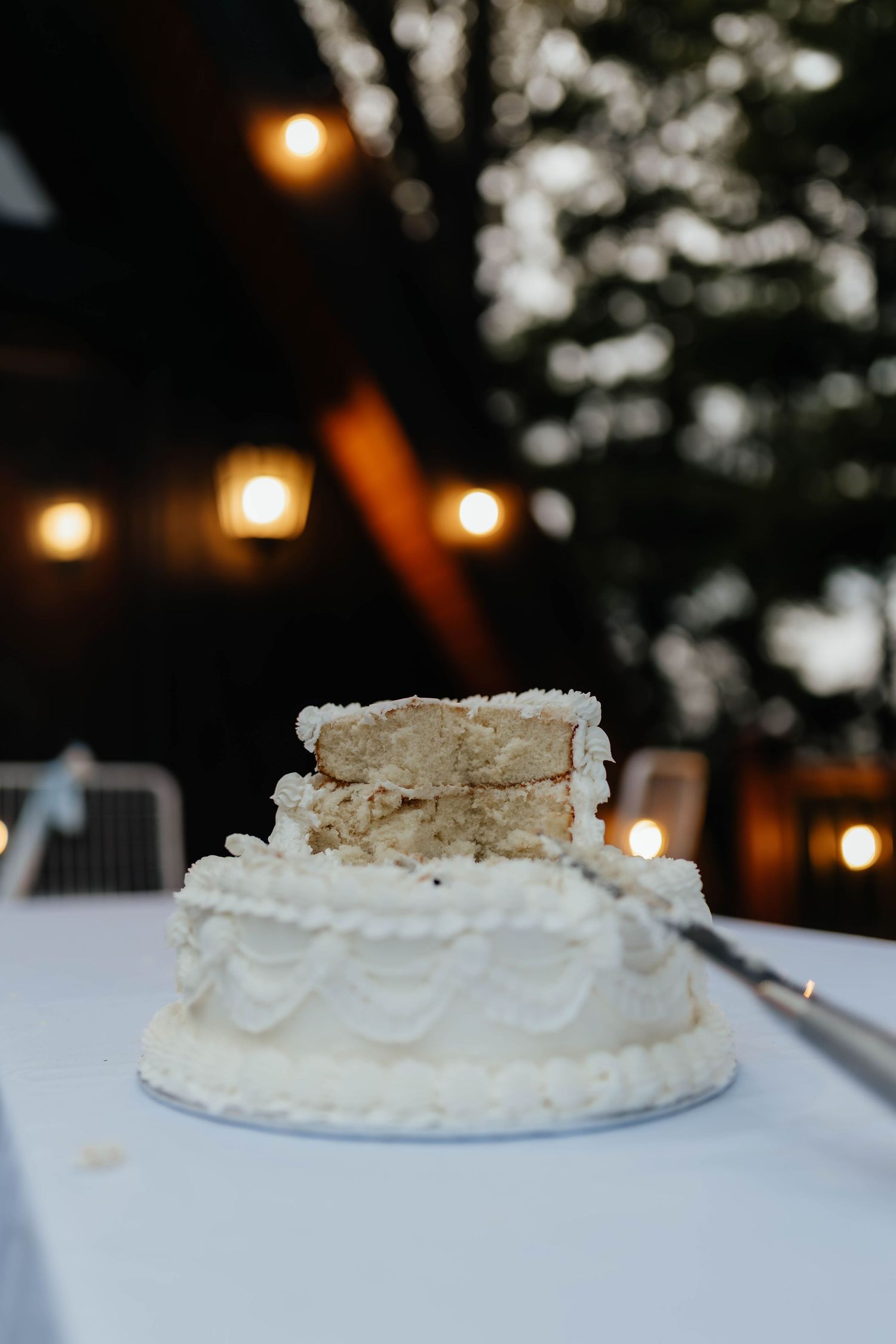 Partially sliced two-tier white wedding cake on a table with string lights in background.
