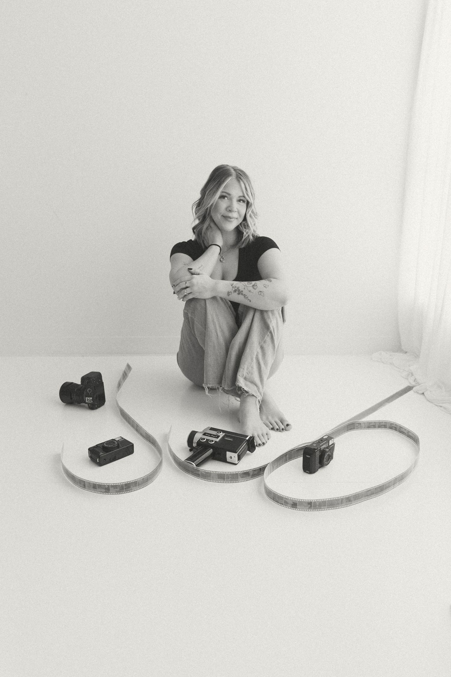 Black and white portrait of a female photographer sitting on floor surrounded by cameras and film.