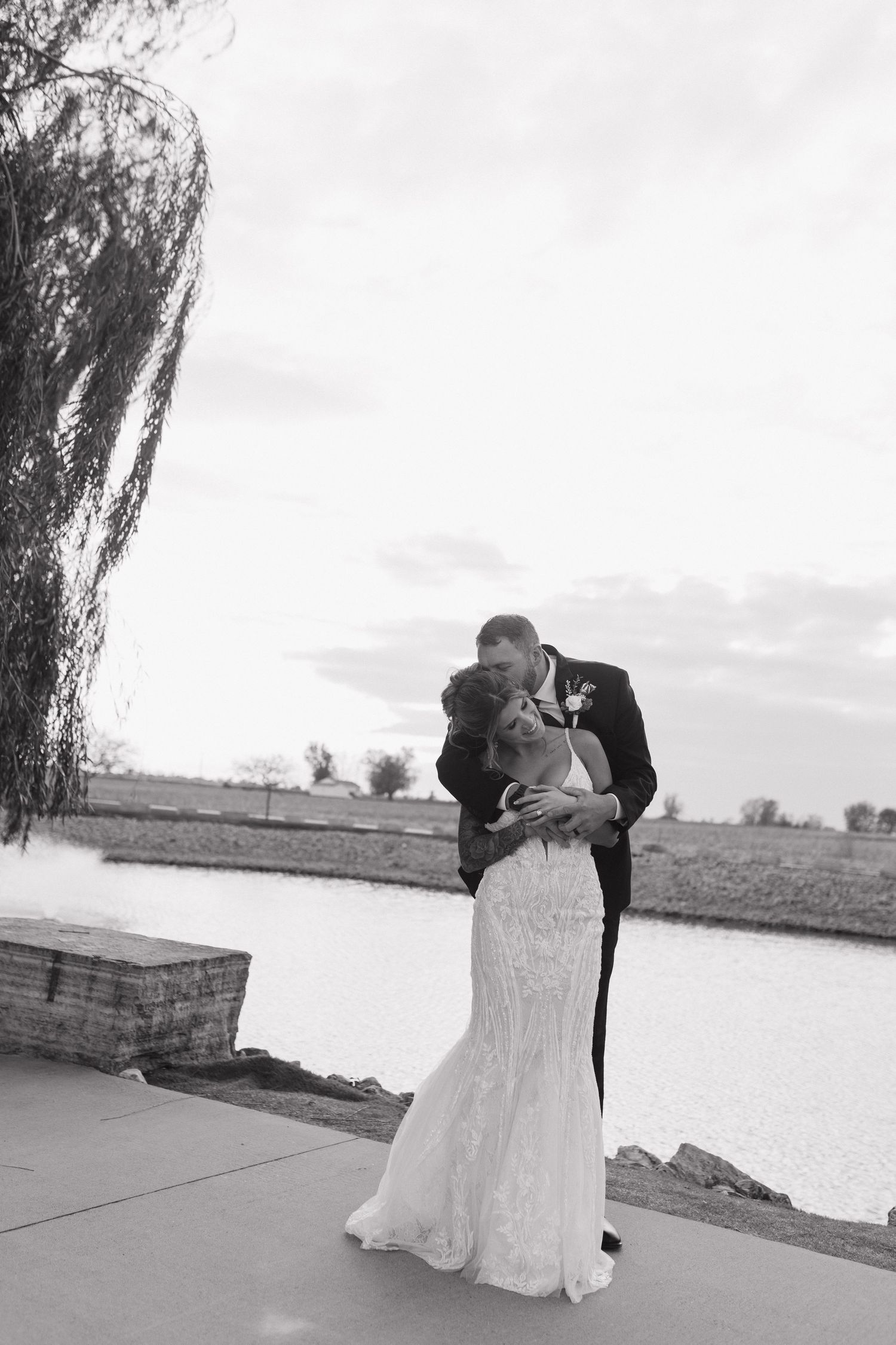 Black and white photo of bride and groom sharing a kiss by a serene pond under cloudy skies.
