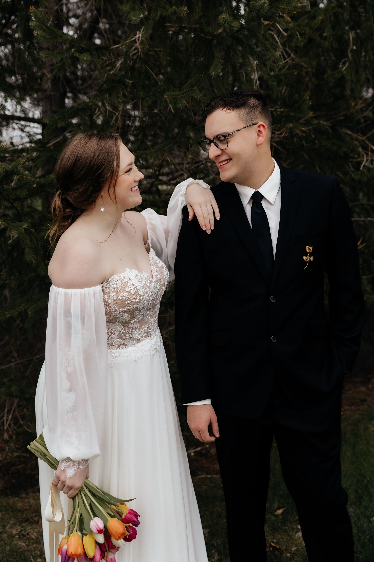 Bride in lace off-shoulder gown holding tulips smiling at groom in black suit beside tall evergreen trees.