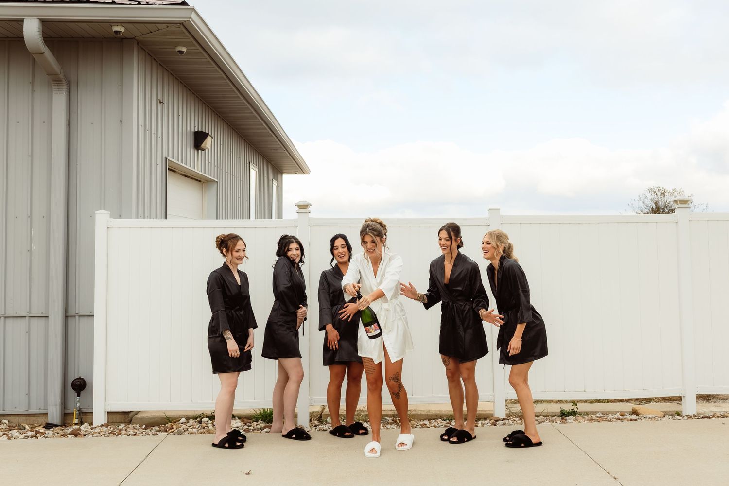 Bride in white robe popping champagne with bridesmaids in matching black robes outdoors.