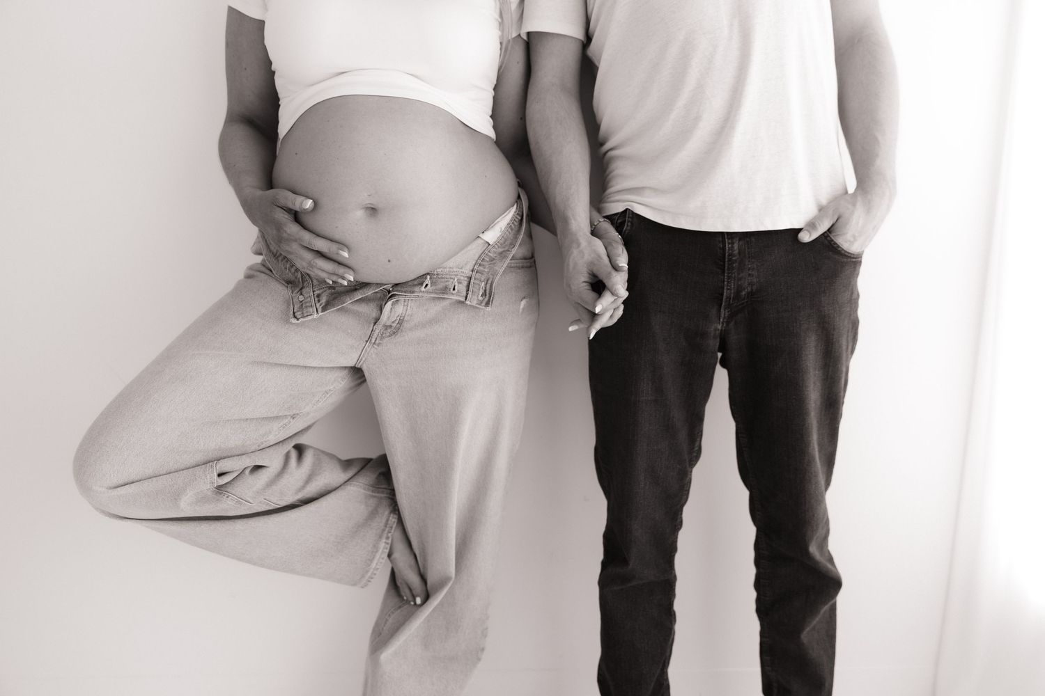 Black and white photo of expecting couple standing together showing pregnant belly in studio.