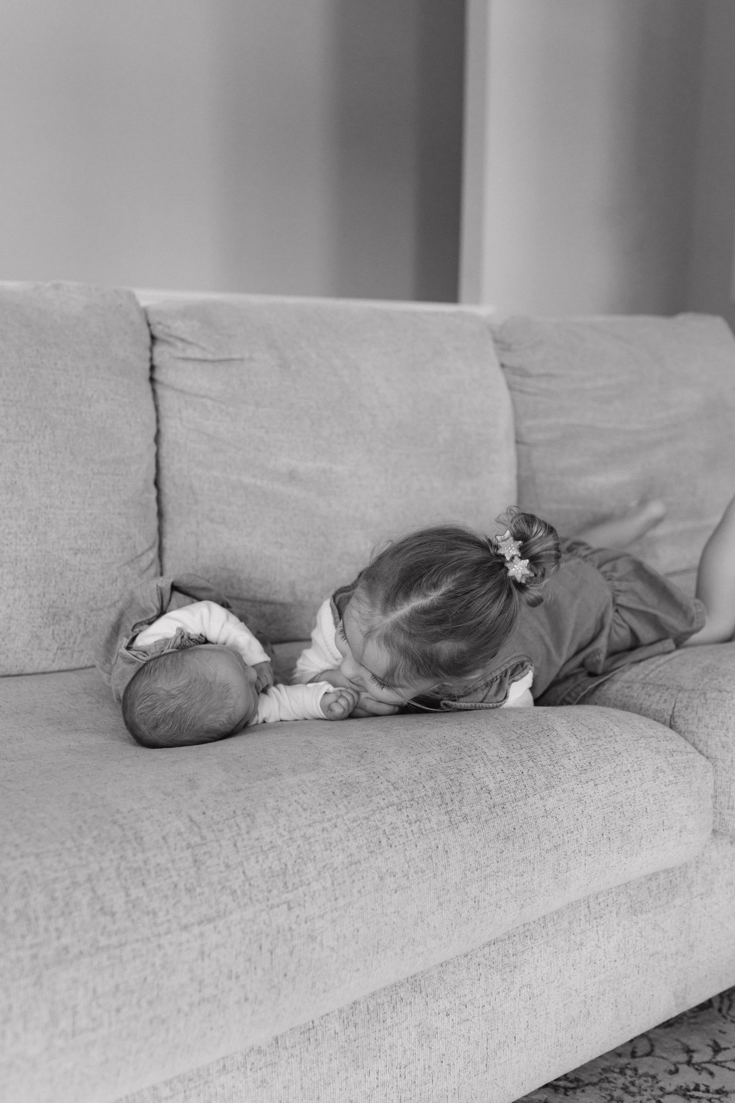 Black and white photo of a young toddler girl leaning over to kiss her newborn sibling on a sofa.