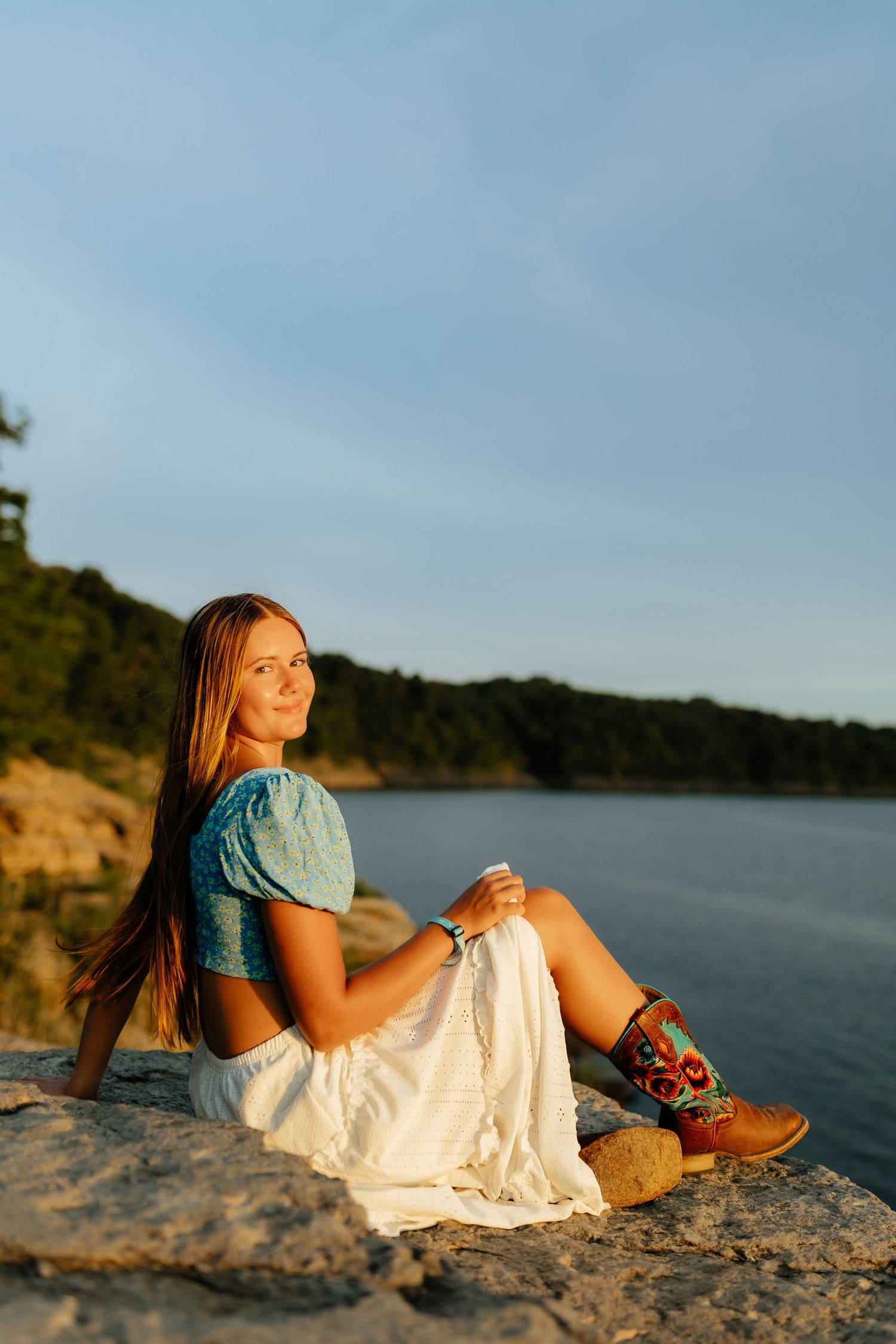 Blonde teen in blue top, white skirt, and cowboy boots sits on rocks by a sunlit lake.