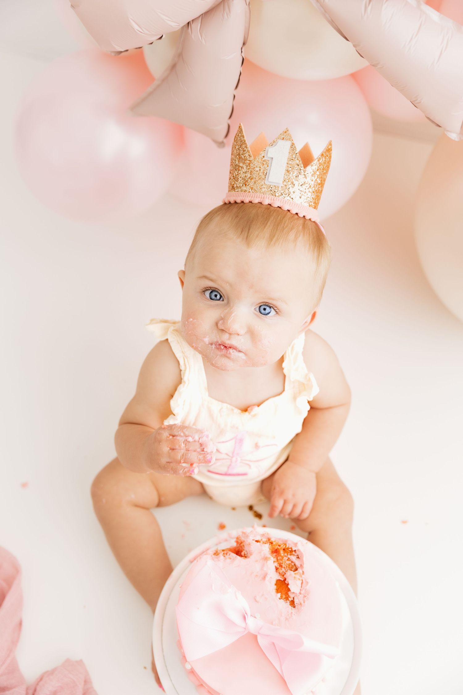 Baby girl in number one crown makes funny face while digging into smashed pink birthday cake.