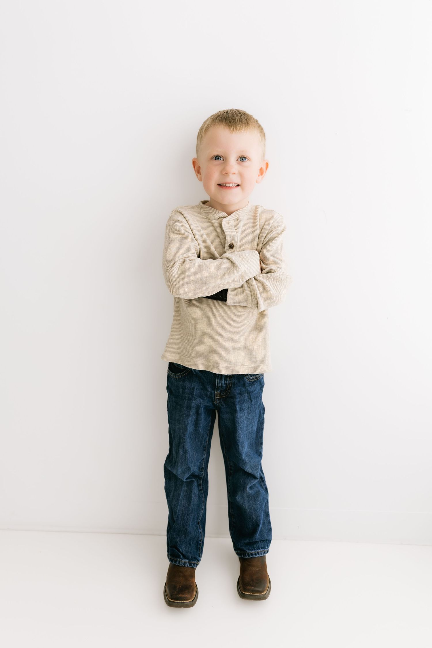Young blonde boy stands with arms crossed smiling confidently in beige henley and jeans against white wall.