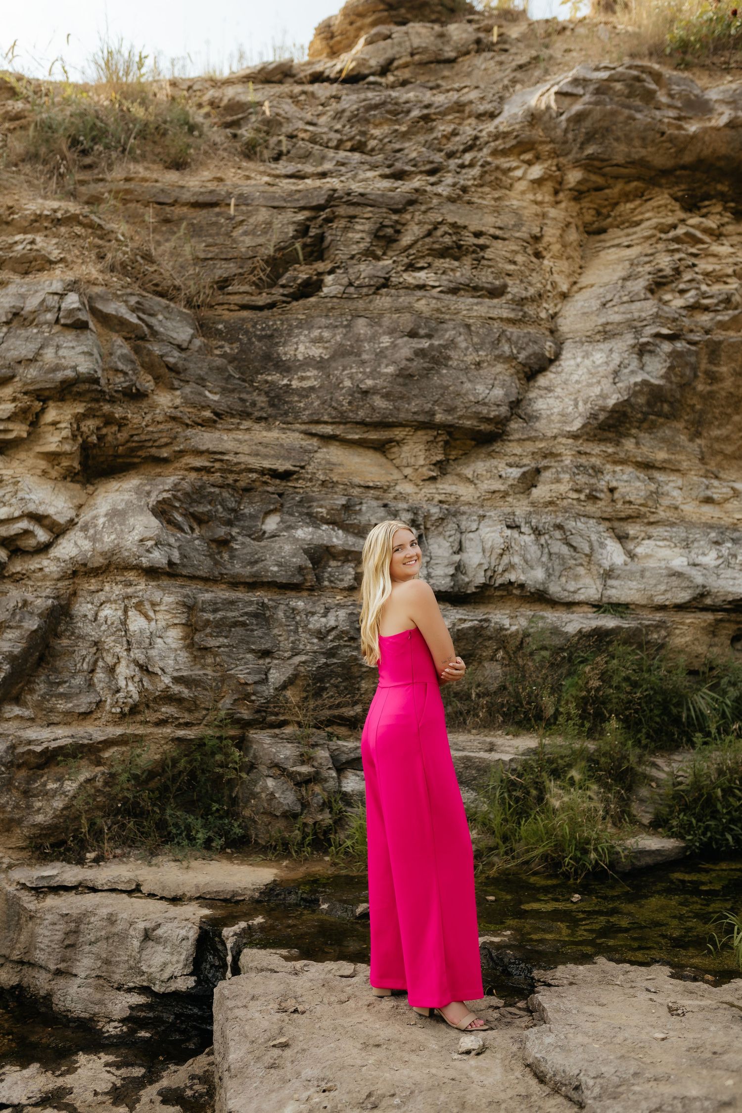 Blonde woman in hot pink strapless maxi dress standing on rocks in front of a rugged stone cliff.
