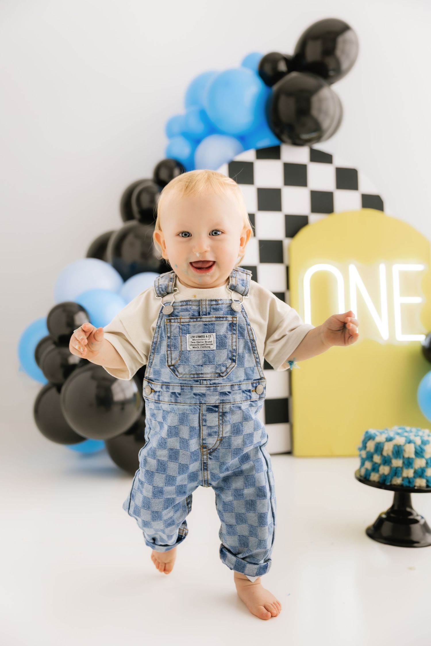 Laughing baby boy in checkered denim overalls celebrating first birthday with race car themed balloon backdrop.