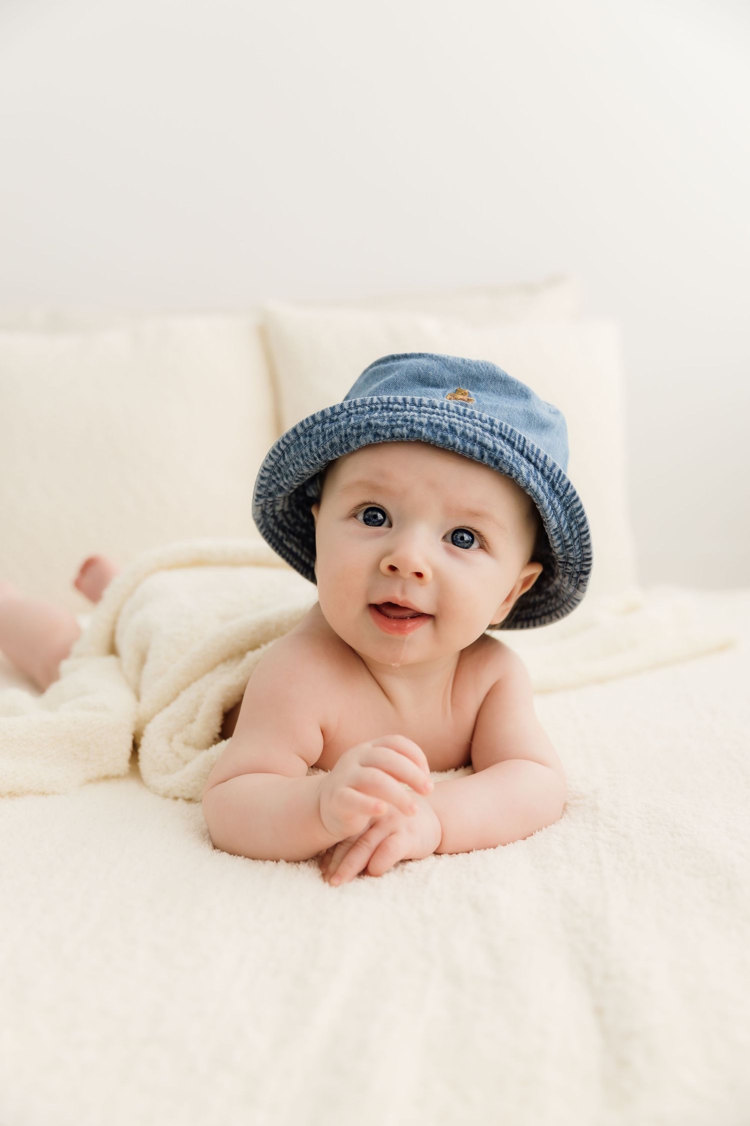 Adorable baby wearing a denim bucket hat lying on a soft cream blanket, smiling curiously at the camera.