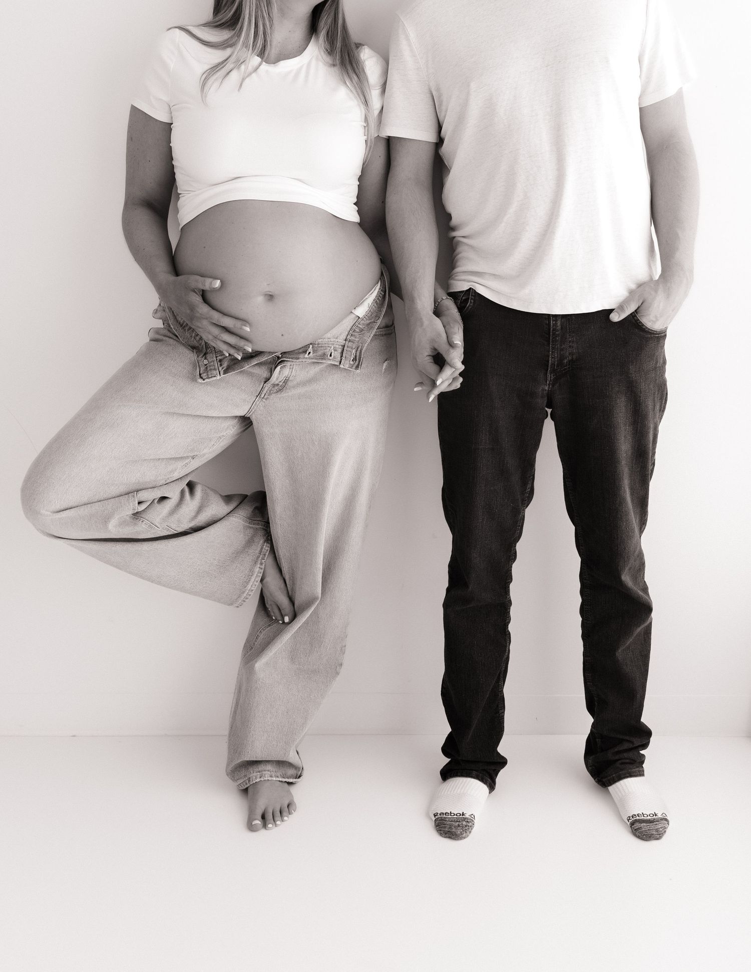 Black and white maternity photo of pregnant woman in white crop top and jeans posing with partner.