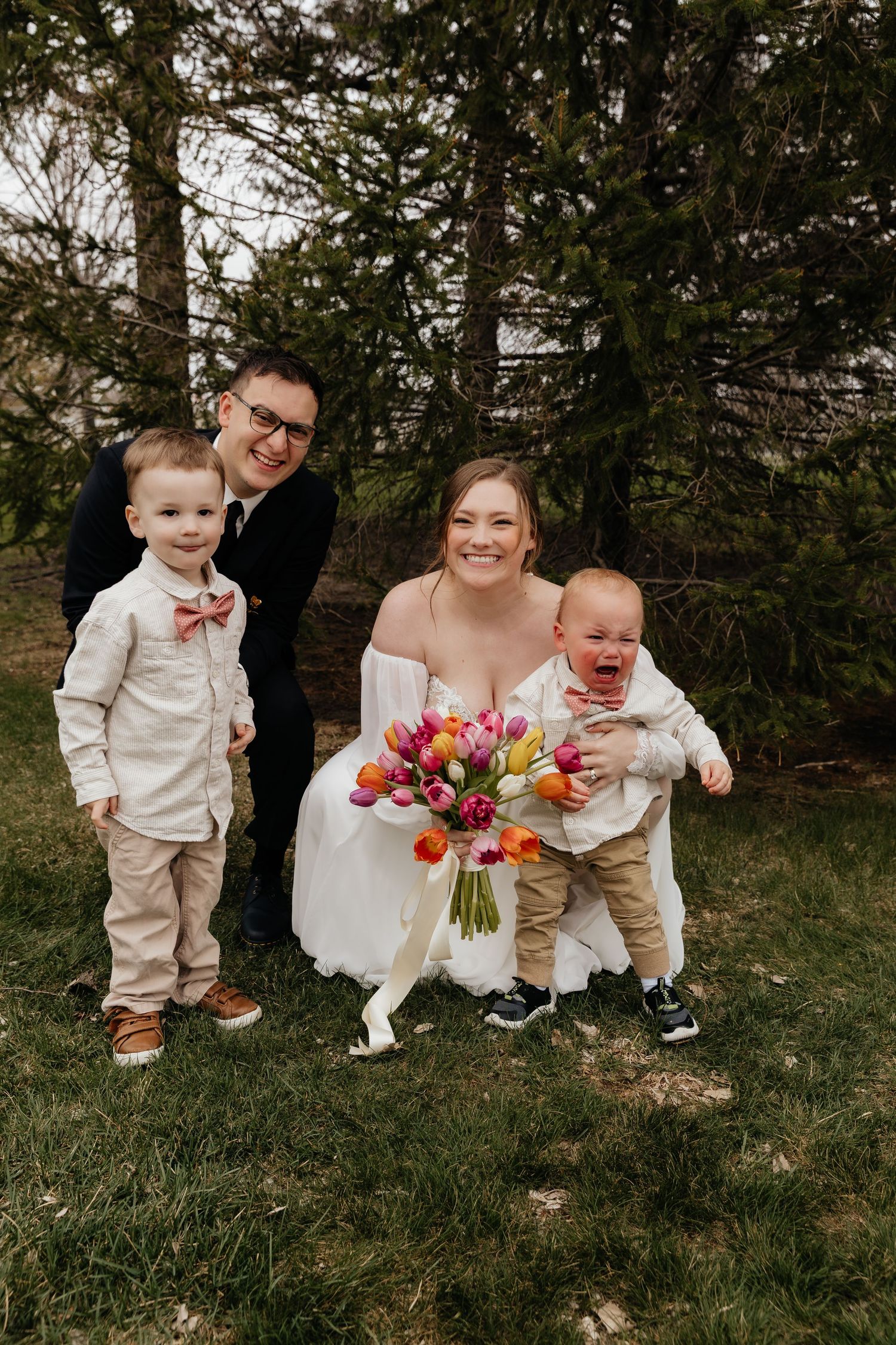 Bride and groom kneel with two young boys holding colorful tulip bouquet outdoors.