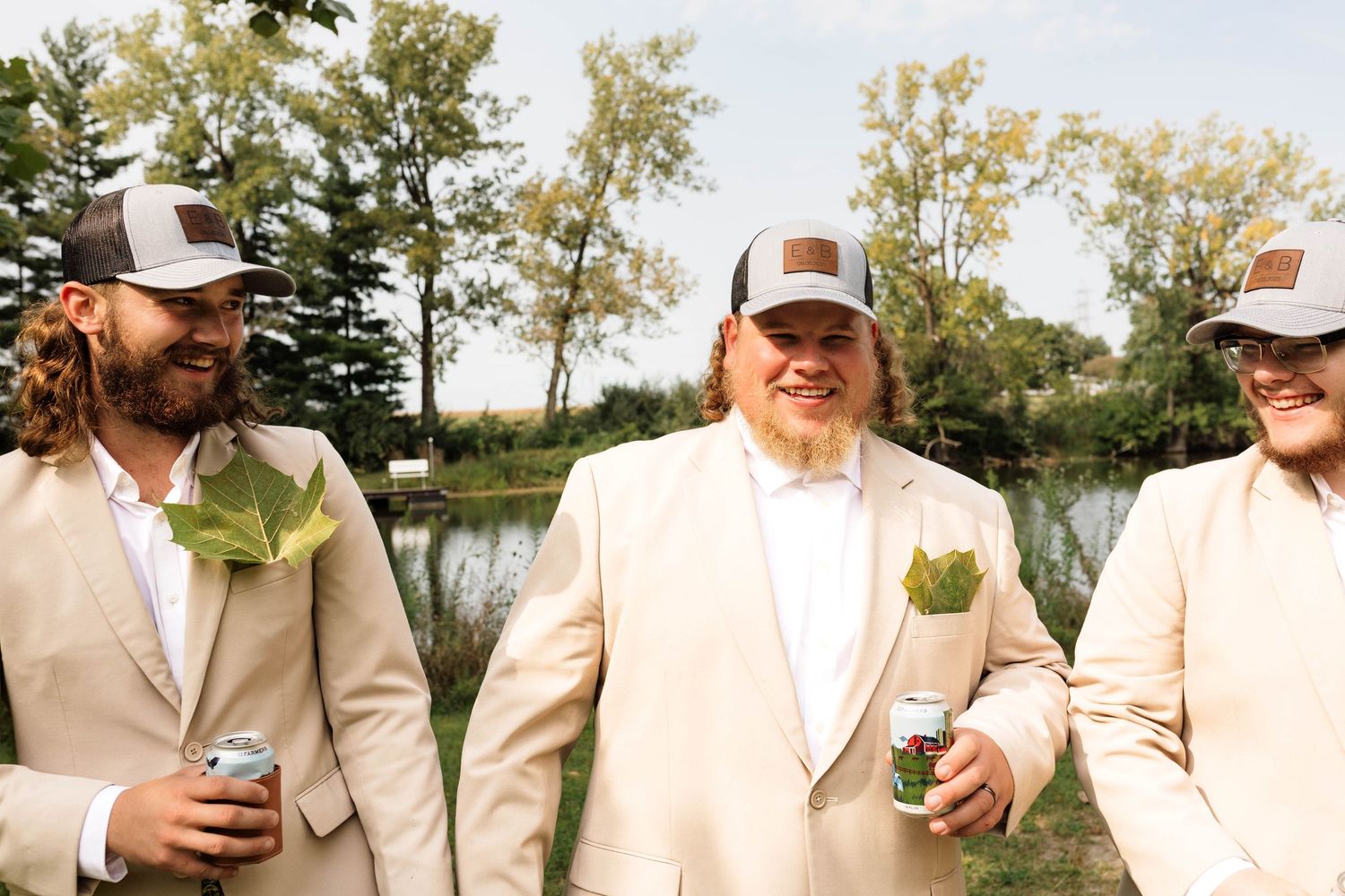 Three groomsmen in tan suits and trucker hats laugh together holding drinks near a scenic pond.