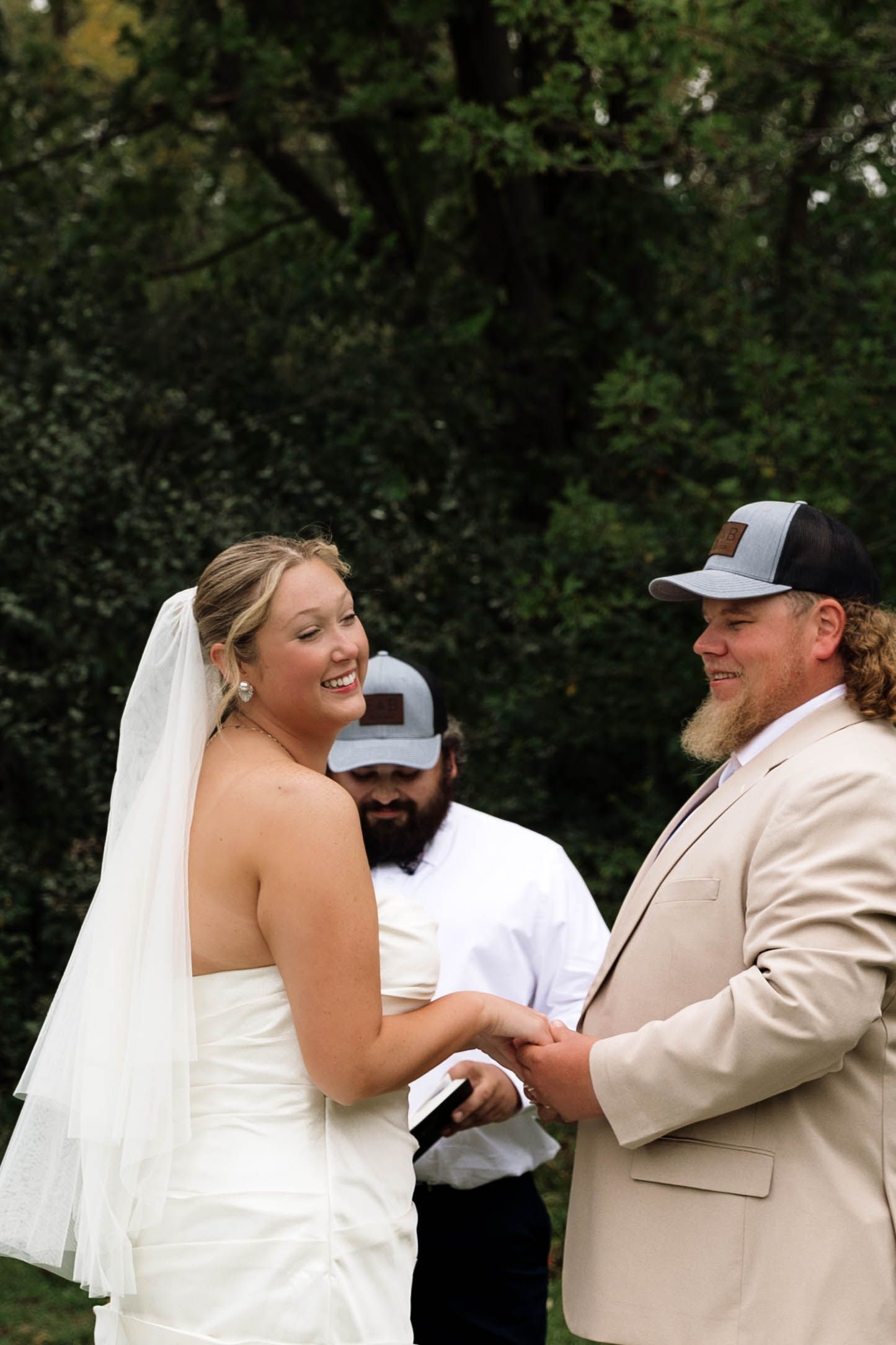 Smiling bride and groom in tan suit and cap exchange vows at outdoor wedding ceremony.