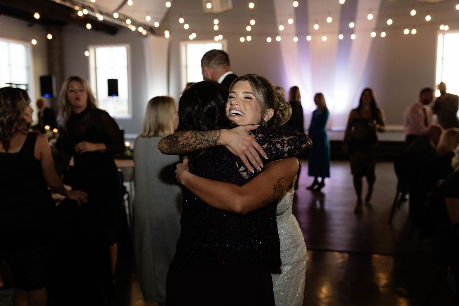 Smiling bride hugs a guest on the dance floor during lively wedding reception with string lights.