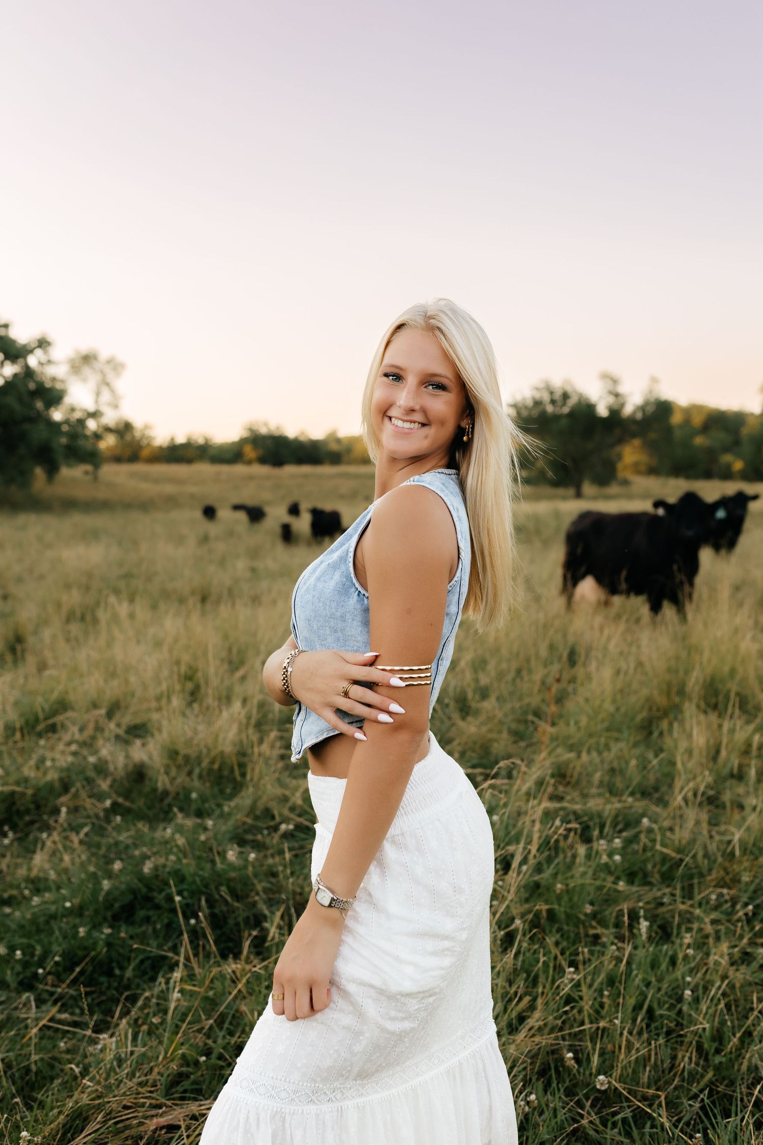 Smiling blonde woman in denim top and white skirt posing in pasture with black cattle.