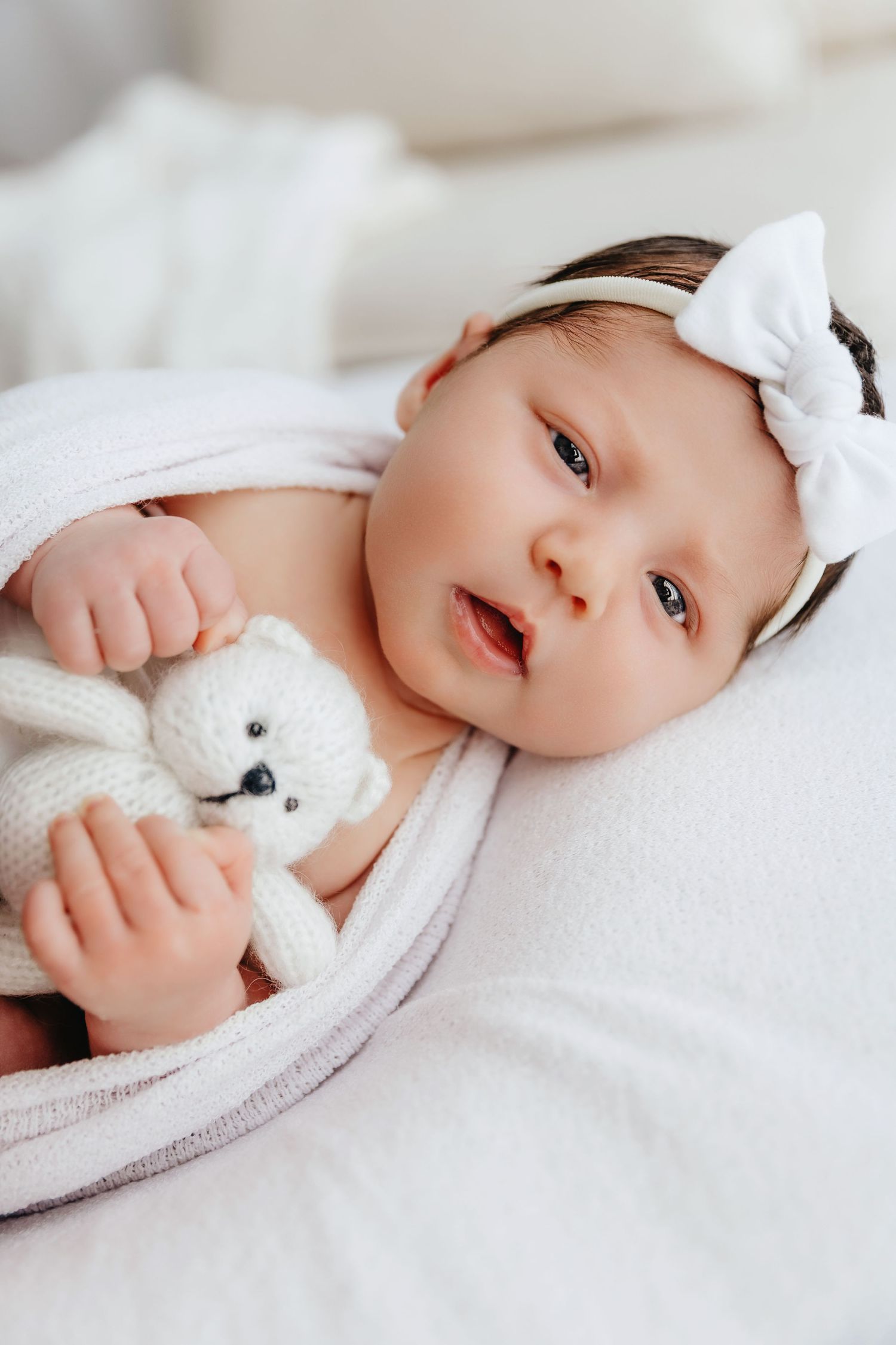 Alert newborn baby with white bow headband holding knitted bunny toy wrapped in pink blanket.
