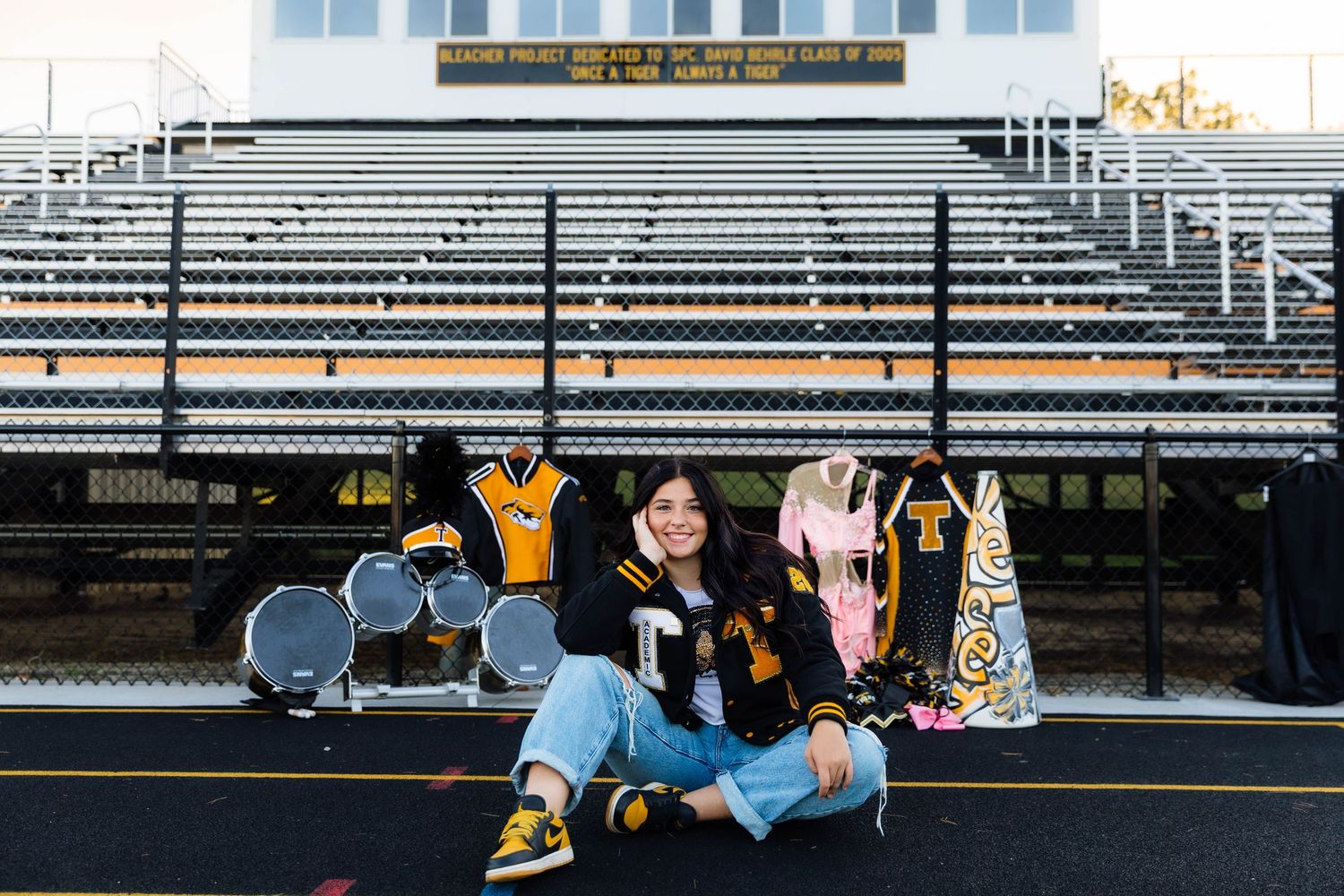 High school senior sits on track in front of bleachers with band and cheer memorabilia displayed.