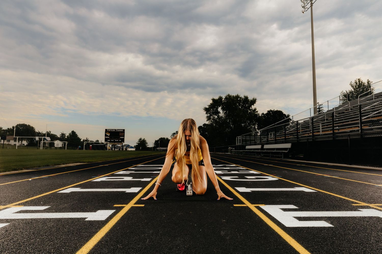 Female track athlete crouches in starting position on school running track under cloudy sky.