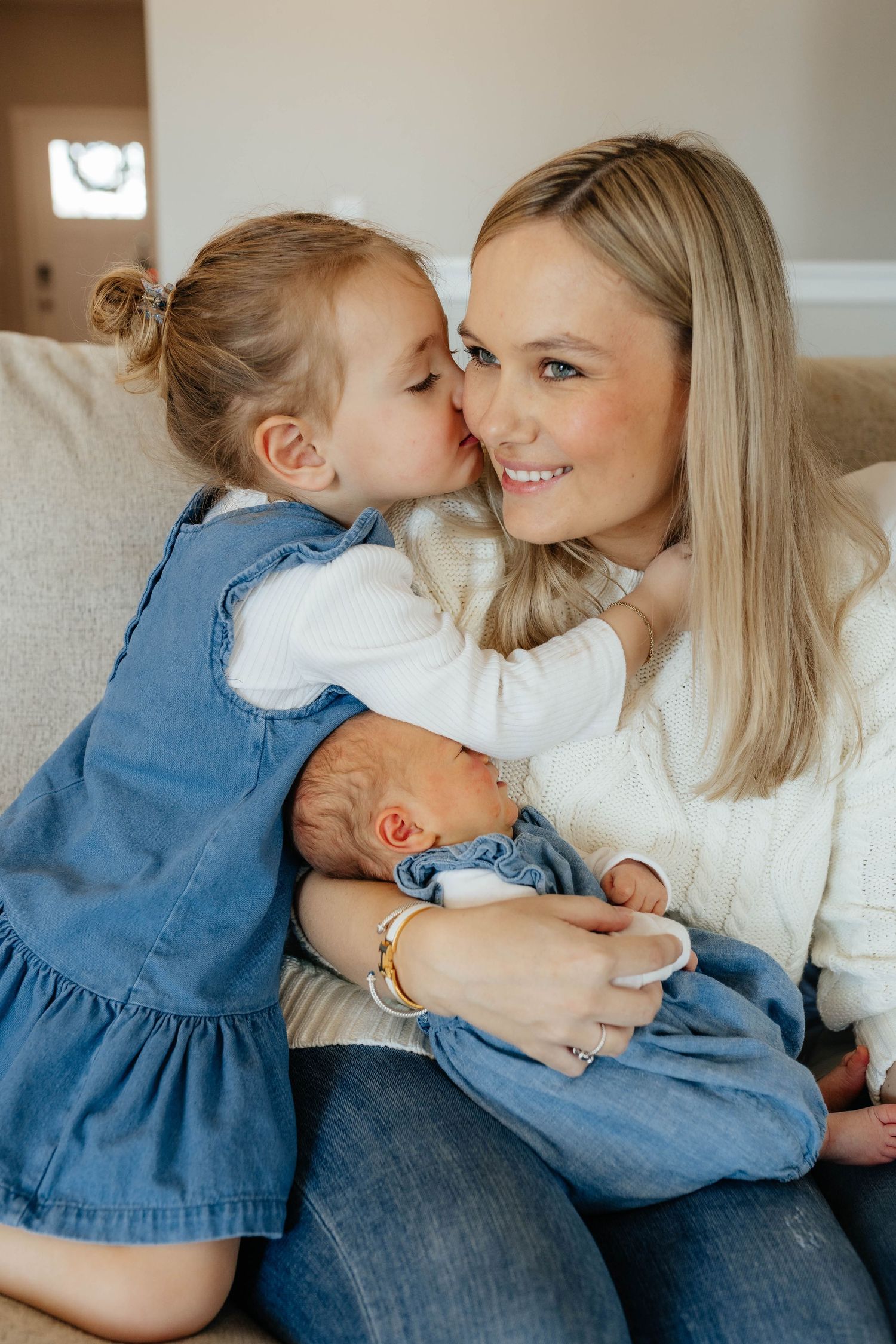 A smiling blonde mother holds her newborn while her toddler daughter in a denim dress gives her a kiss on the cheek.