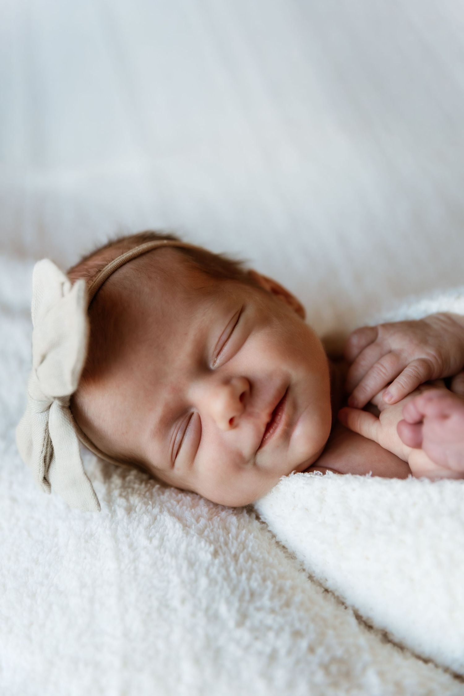 Peaceful sleeping newborn baby girl with cream bow headband wrapped in white blanket.