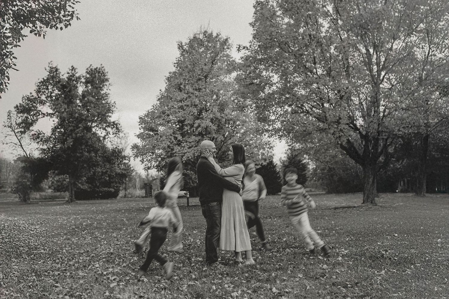 Black and white photo of family hugging together in a circle in a park surrounded by trees.