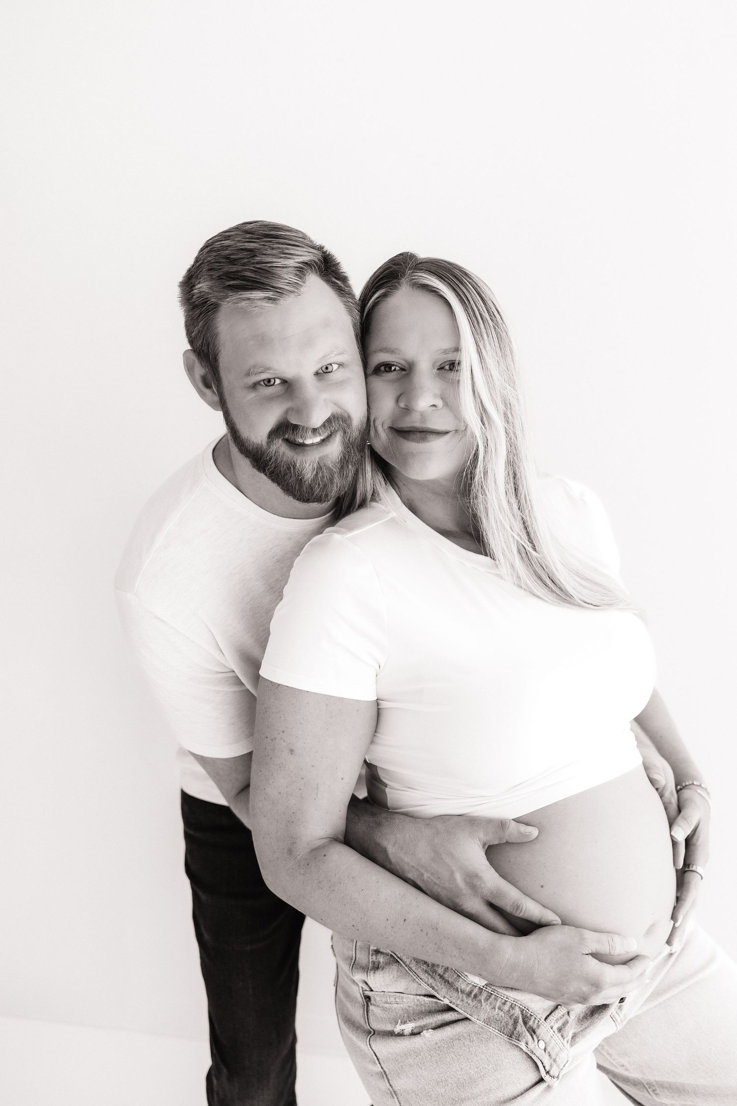Black and white portrait of smiling expecting couple embracing pregnant belly in studio.