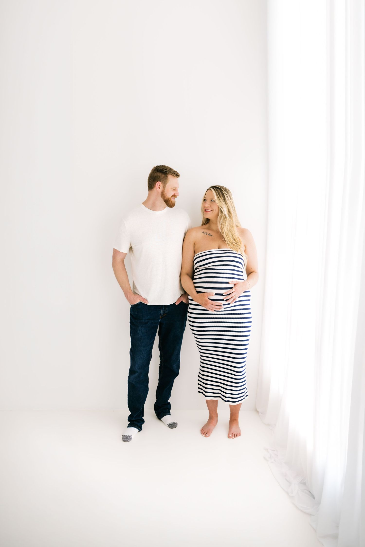Expecting couple standing together in bright white studio, woman in striped strapless maternity dress, man in white tee.