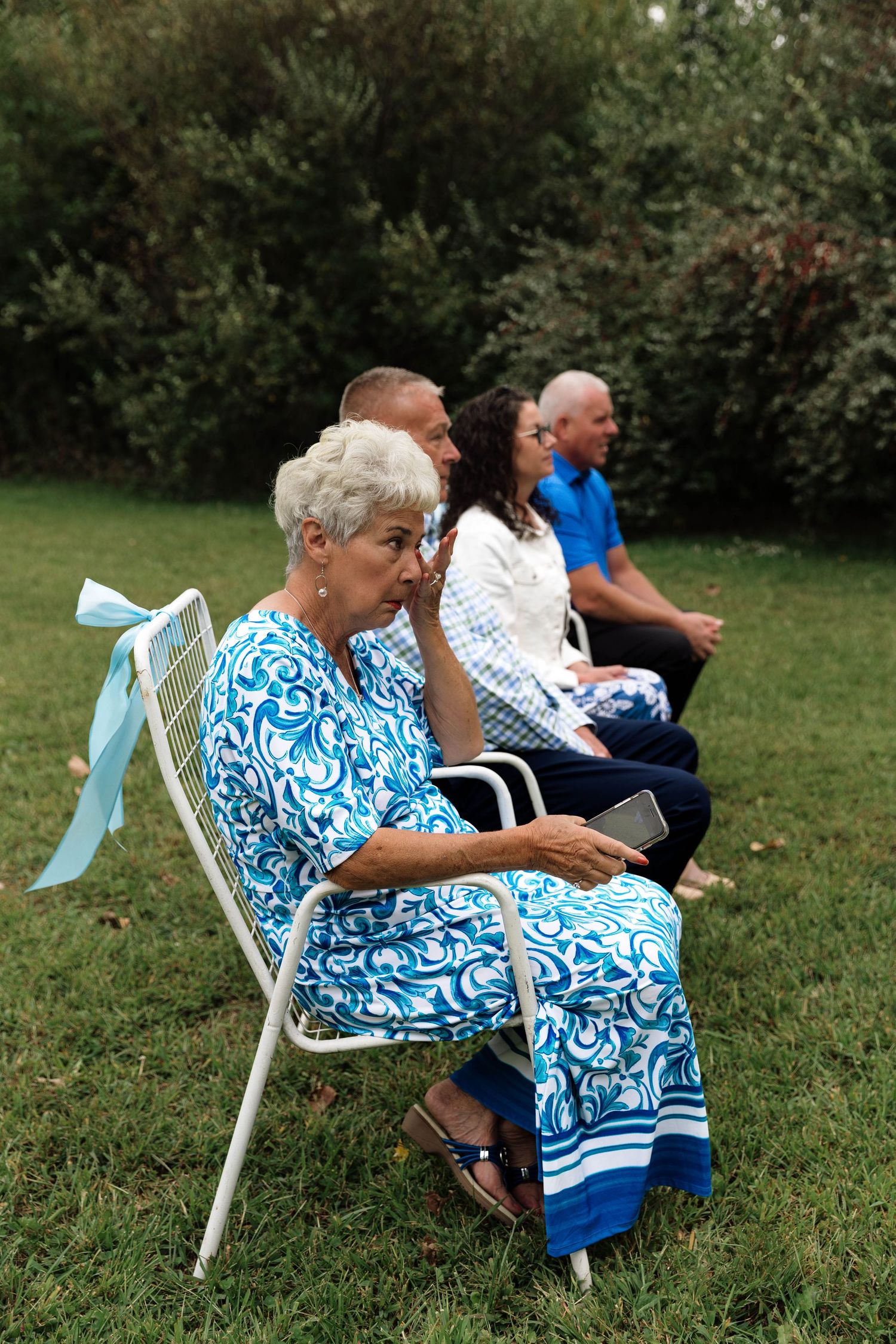 Elderly woman in blue floral dress wipes tears while seated at outdoor wedding ceremony.