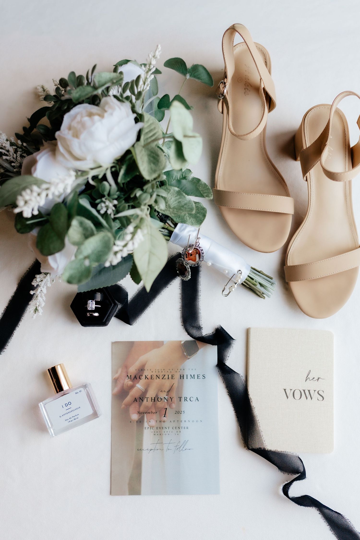 Bridal flat lay featuring bouquet, nude sandals, vow book, perfume, and wedding invitation on white.