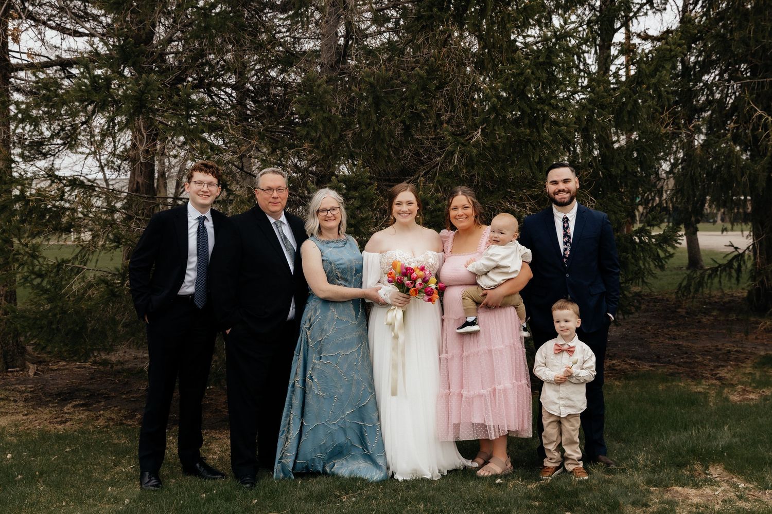 Wedding party family portrait with bride, groom, and relatives posing outdoors near trees.