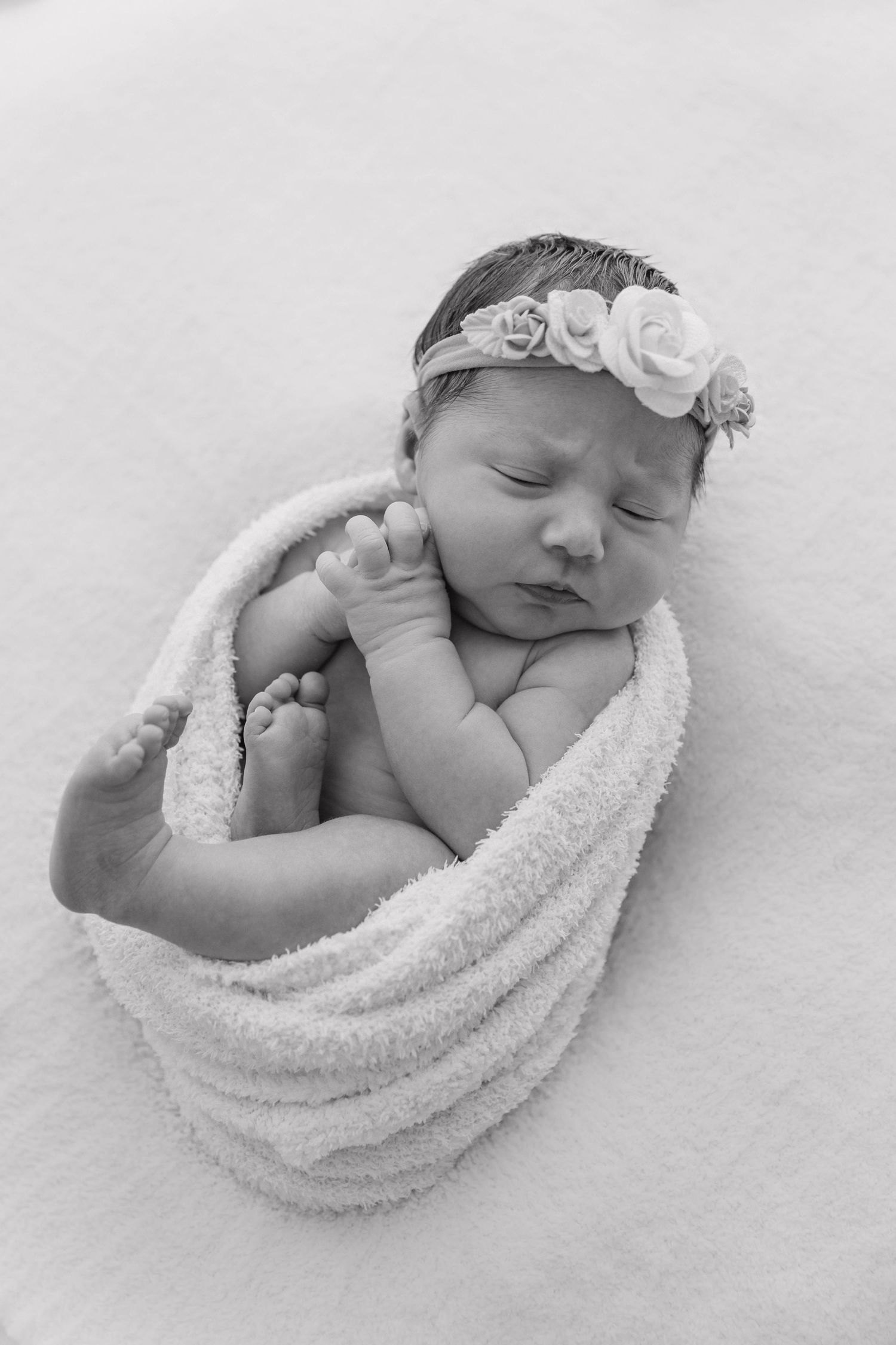 Black and white portrait of swaddled newborn baby with floral headband sleeping peacefully.