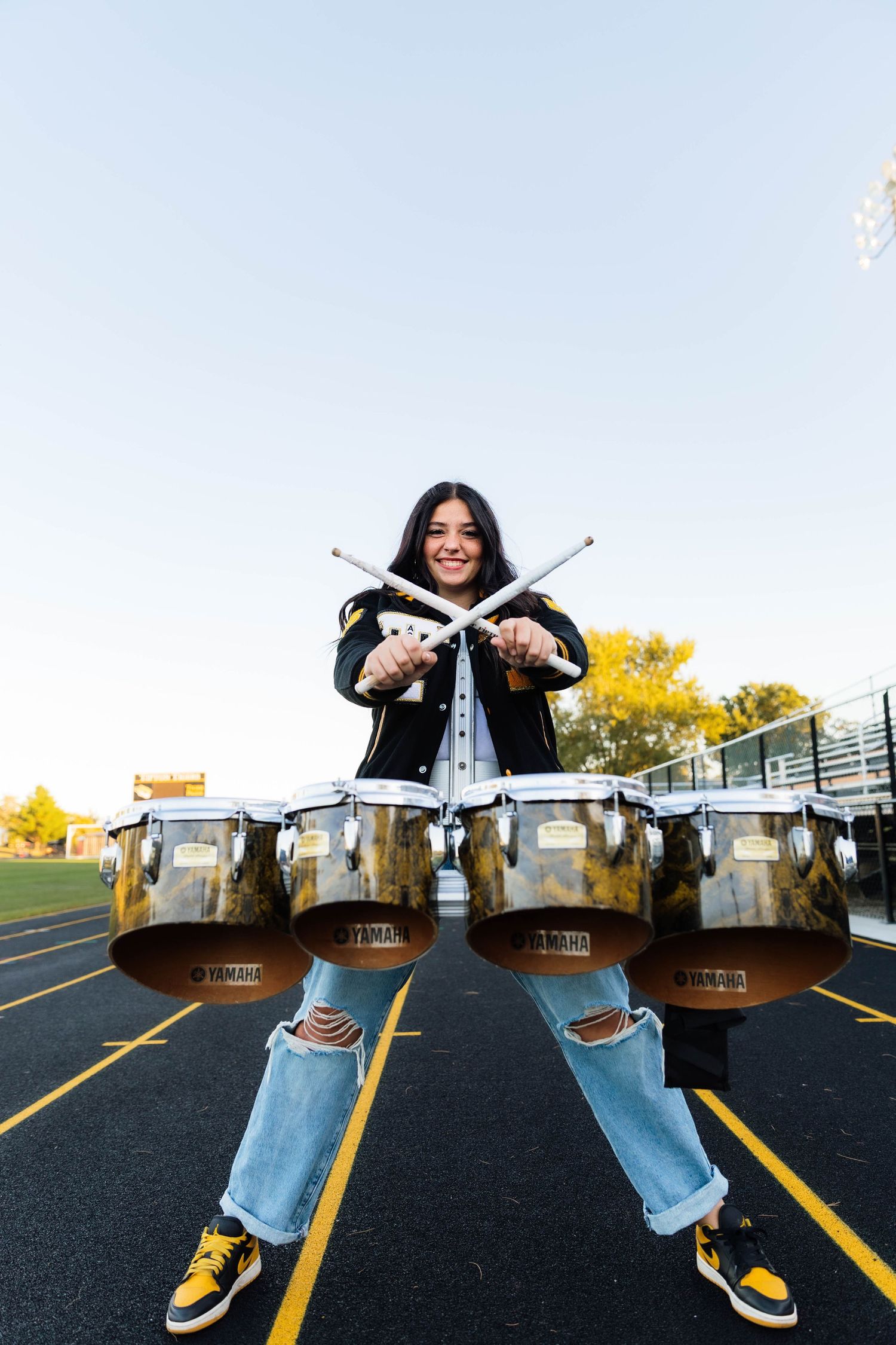 High school drummer in varsity jacket plays Yamaha tenor drums on colorful track at dusk.