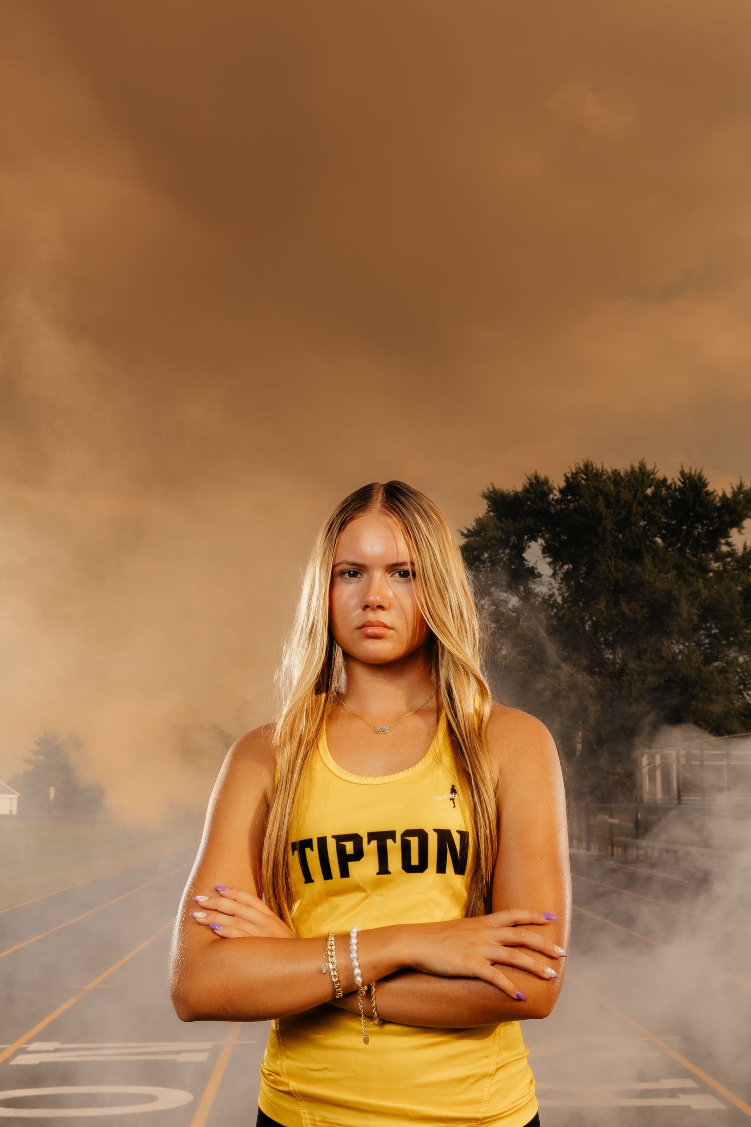 Confident blonde female athlete in Tipton jersey stands arms crossed amid dramatic smoky backdrop.