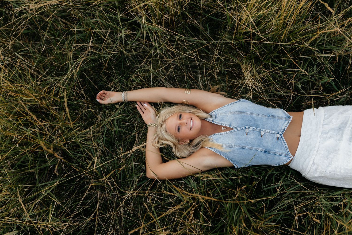Smiling blonde teen girl lying in tall green grass wearing denim overalls during golden hour.