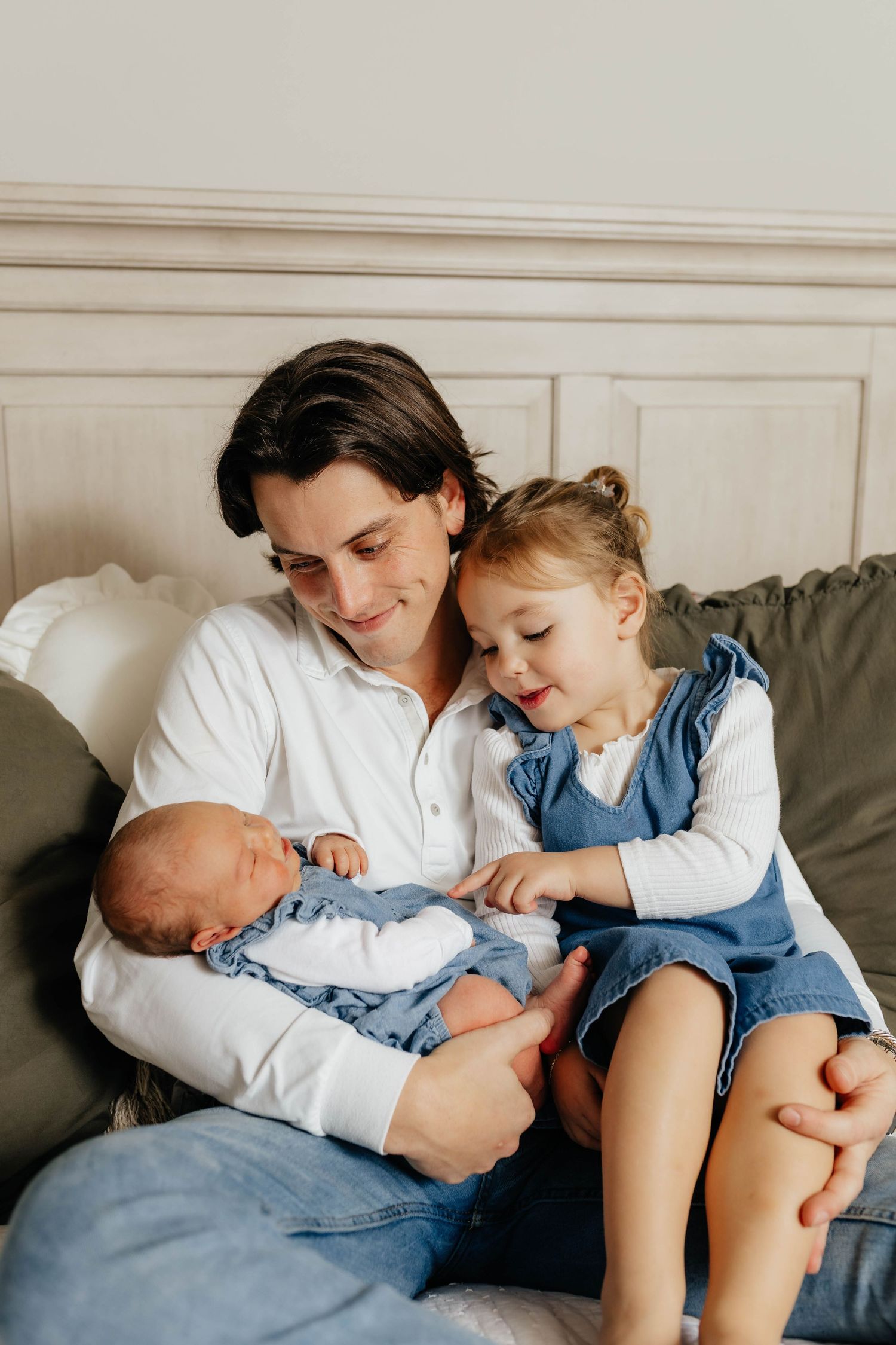 A dark-haired father sits on a bed holding his newborn while his young daughter in a denim dress looks on adoringly.