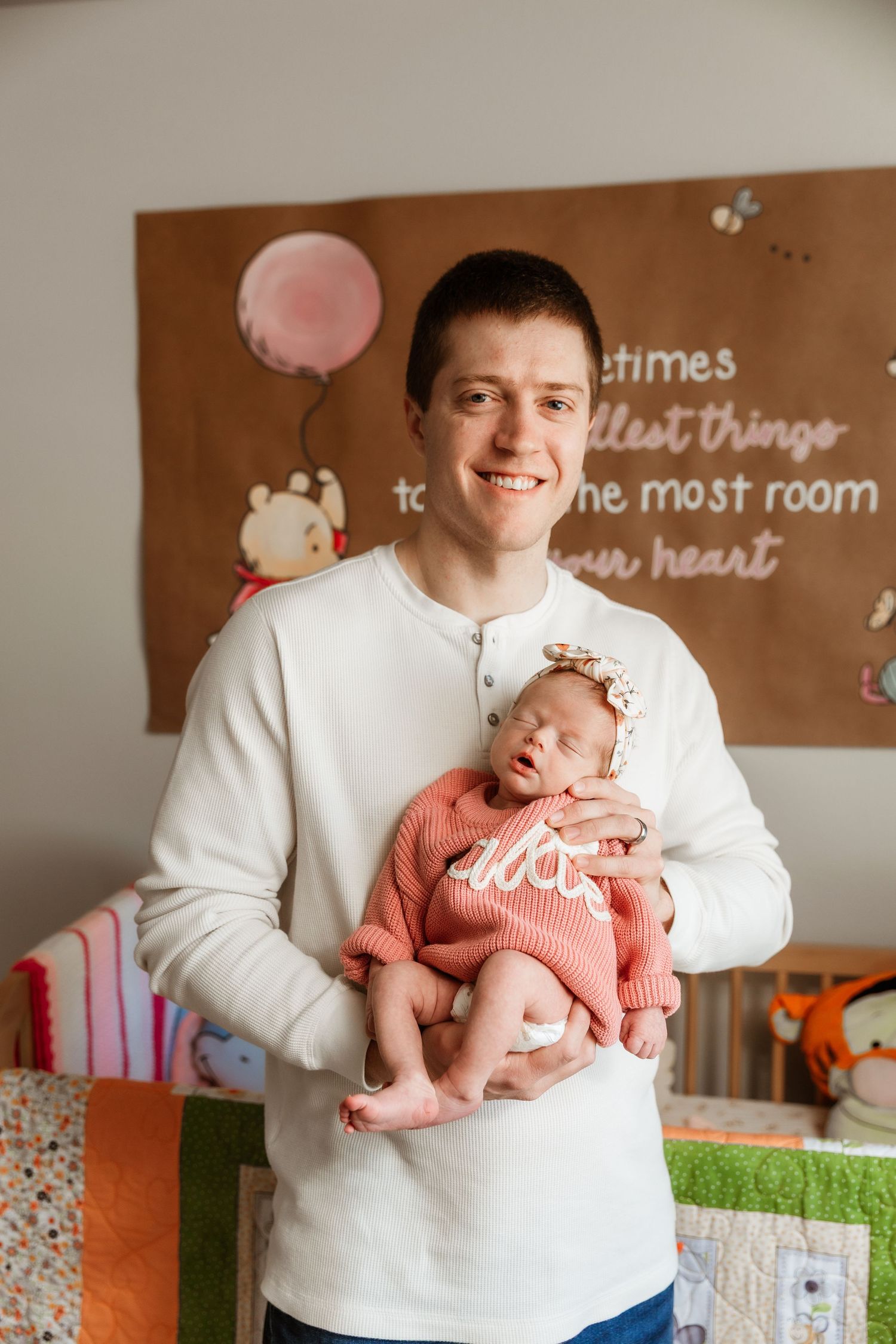 Smiling father in white shirt holds his newborn baby girl in pink outfit in nursery.