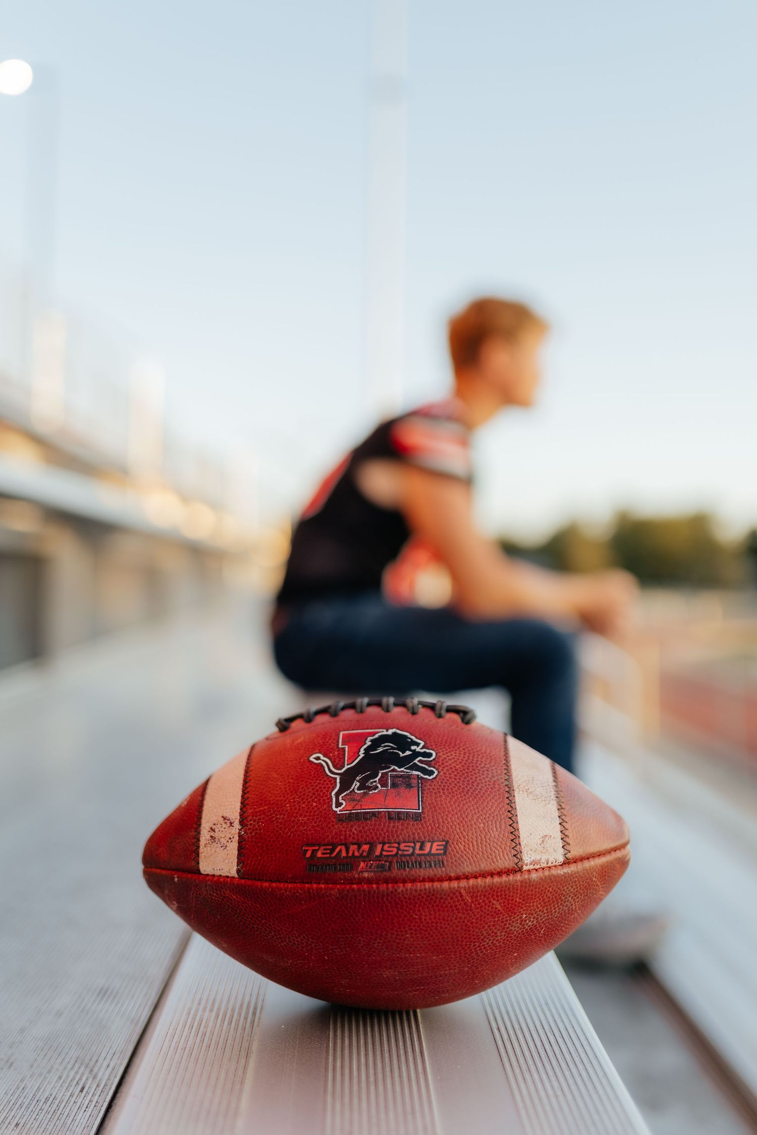 Close-up of a Lions Team Issue football on bleachers with player blurred in background.