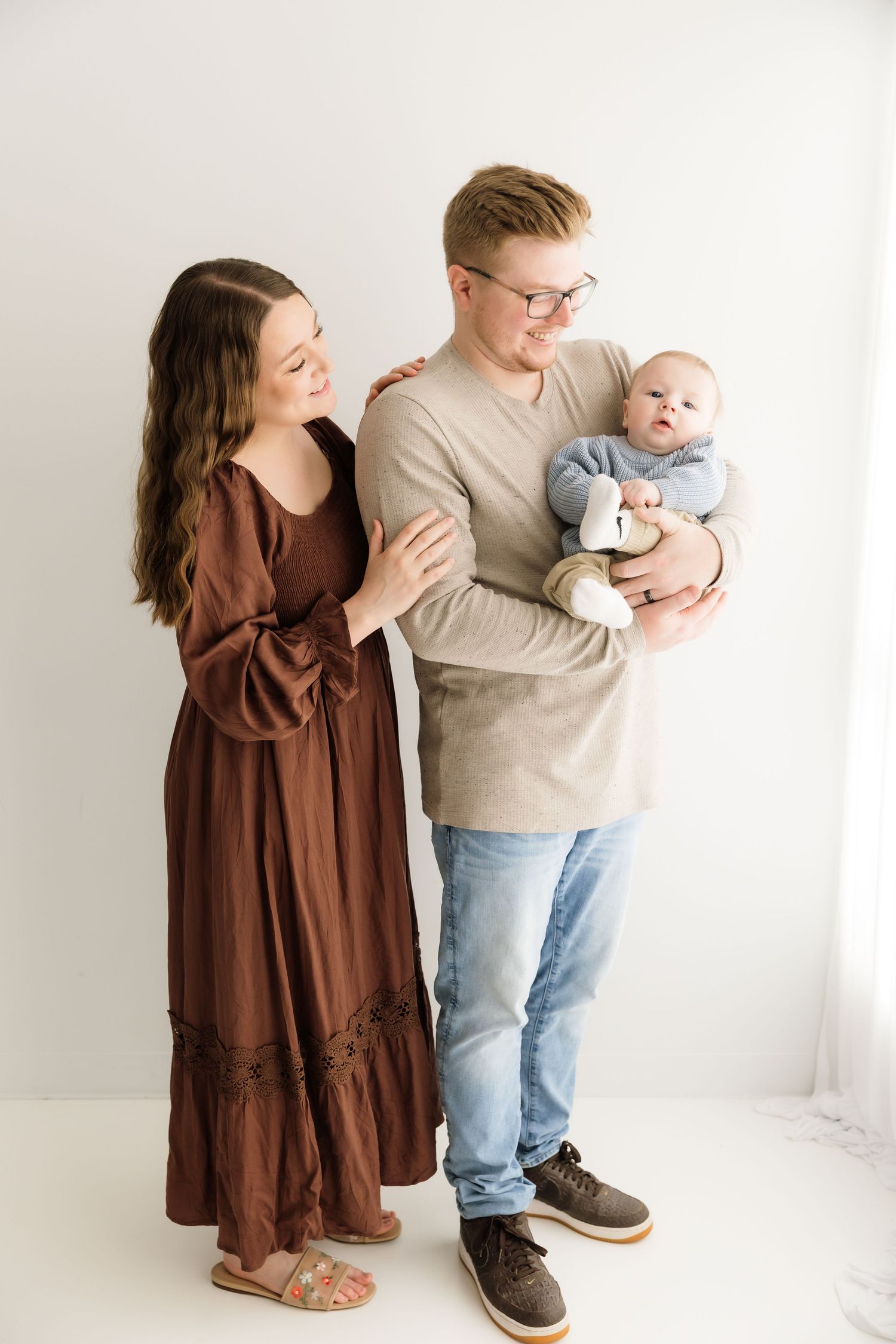 Young family with mother in brown dress and father holding baby in blue sweater.