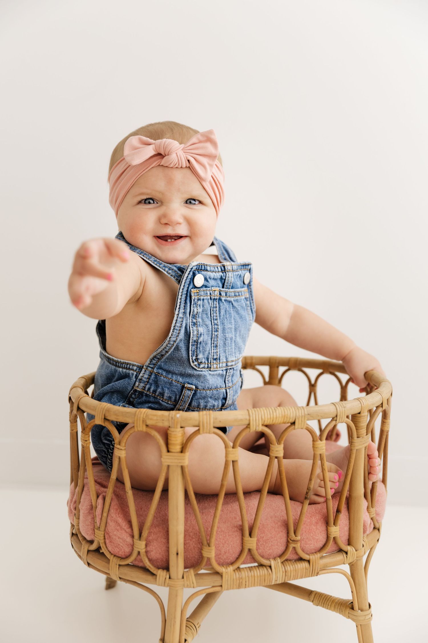 Smiling baby girl in denim overalls and pink bow headband sitting in rattan basket reaching forward.