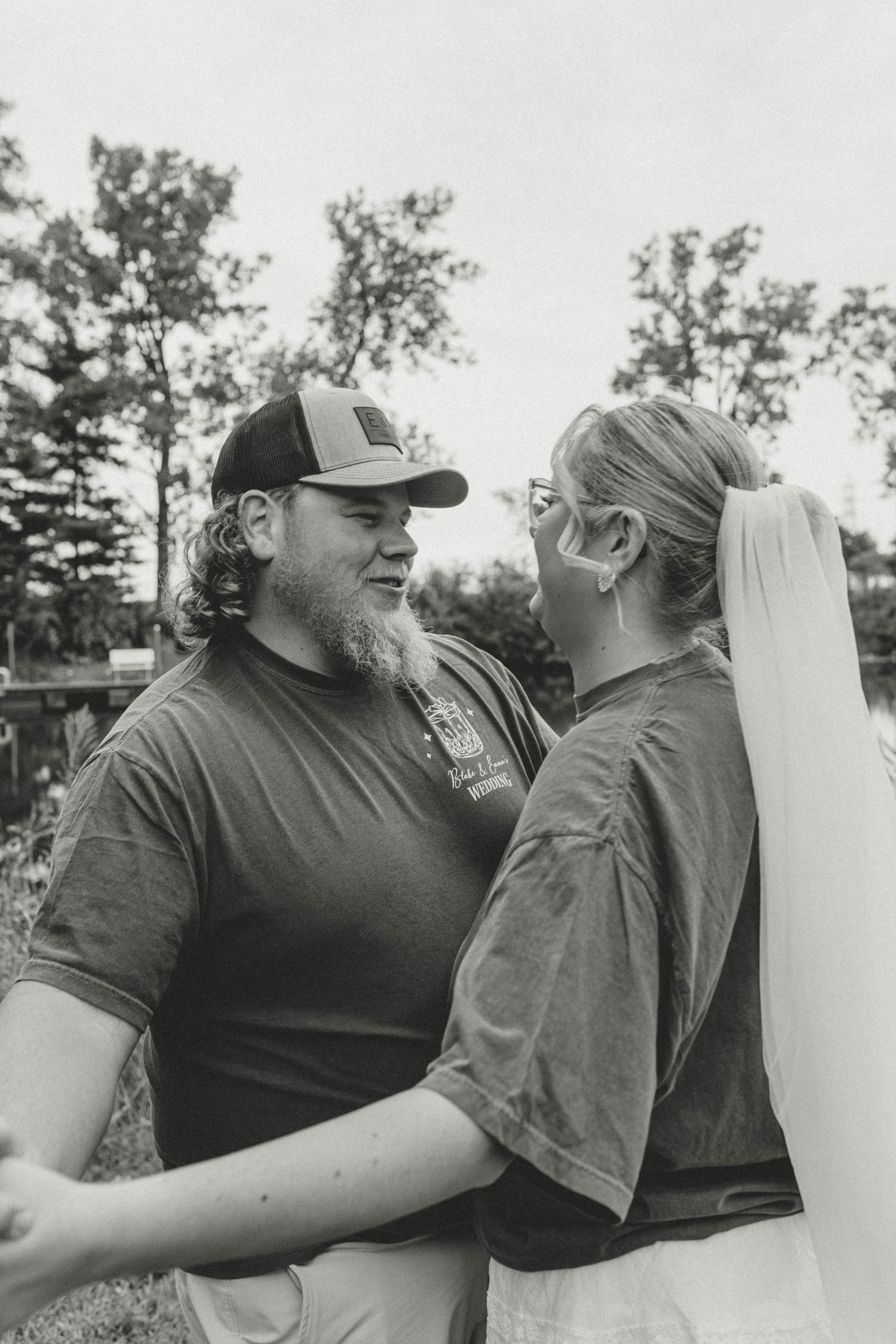 Black and white photo of a bearded groom in a cap embracing his bride outdoors.