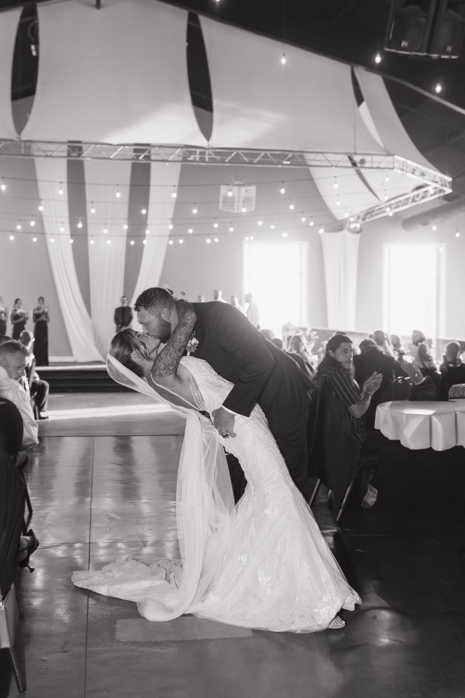 Bride and groom share romantic dip kiss on dance floor surrounded by wedding guests.