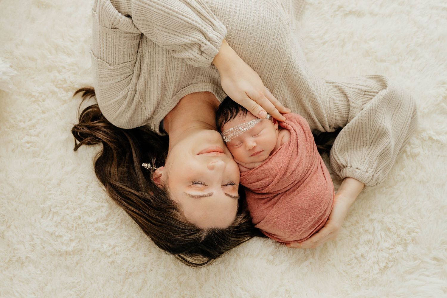 Overhead view of mother and pink-swaddled newborn with sparkle headband lying together on white rug.