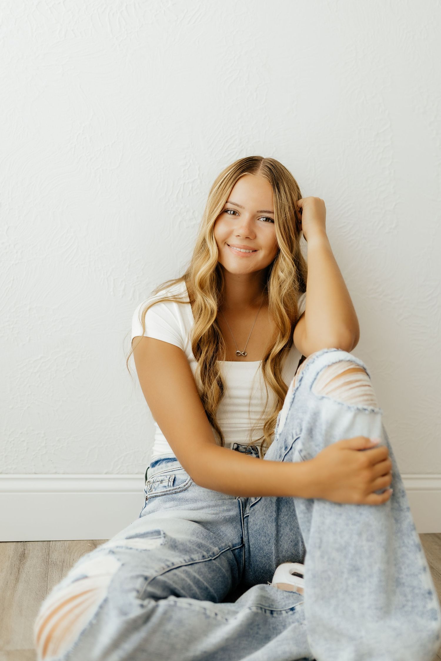 Smiling blonde teenage girl in white top and ripped jeans sits against white wall indoors.