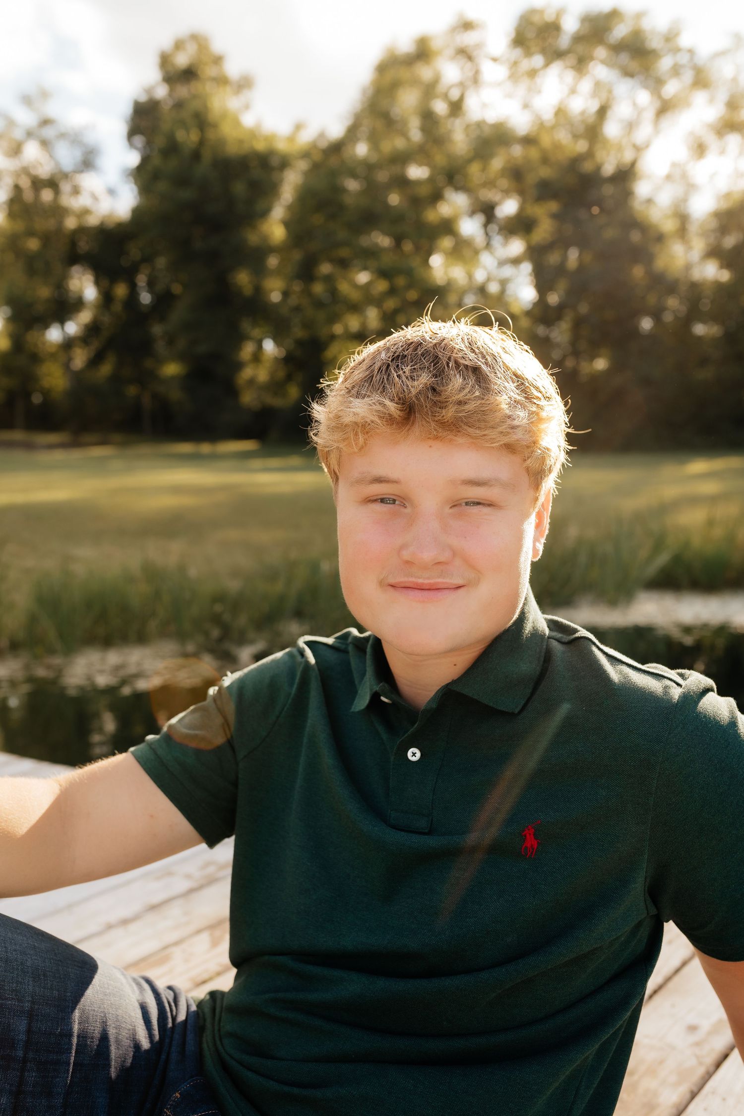 Smiling teenage boy in green polo shirt sitting outdoors by a pond in golden sunlight.