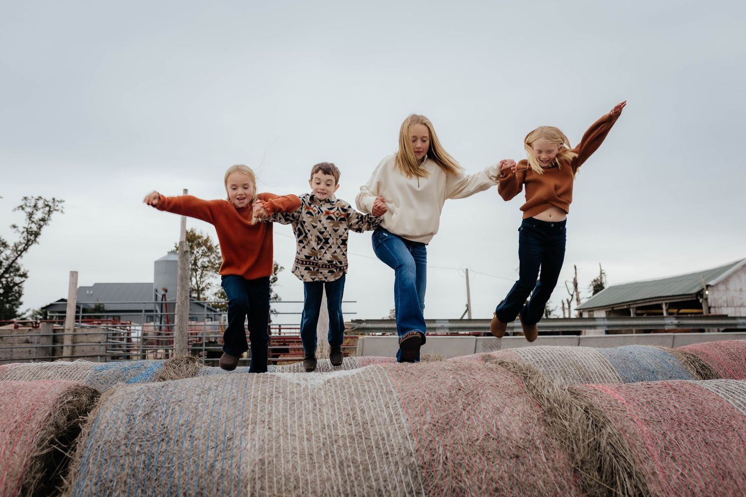 Four children joyfully jumping together on a large hay bale under an overcast sky.