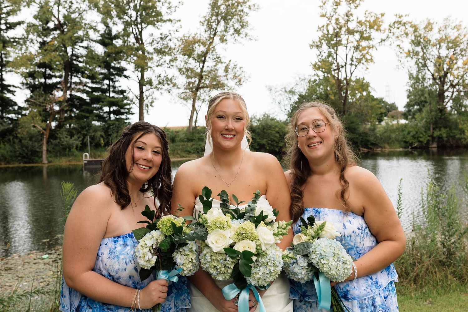 Bride with two bridesmaids in blue floral dresses holding white bouquets by a pond.